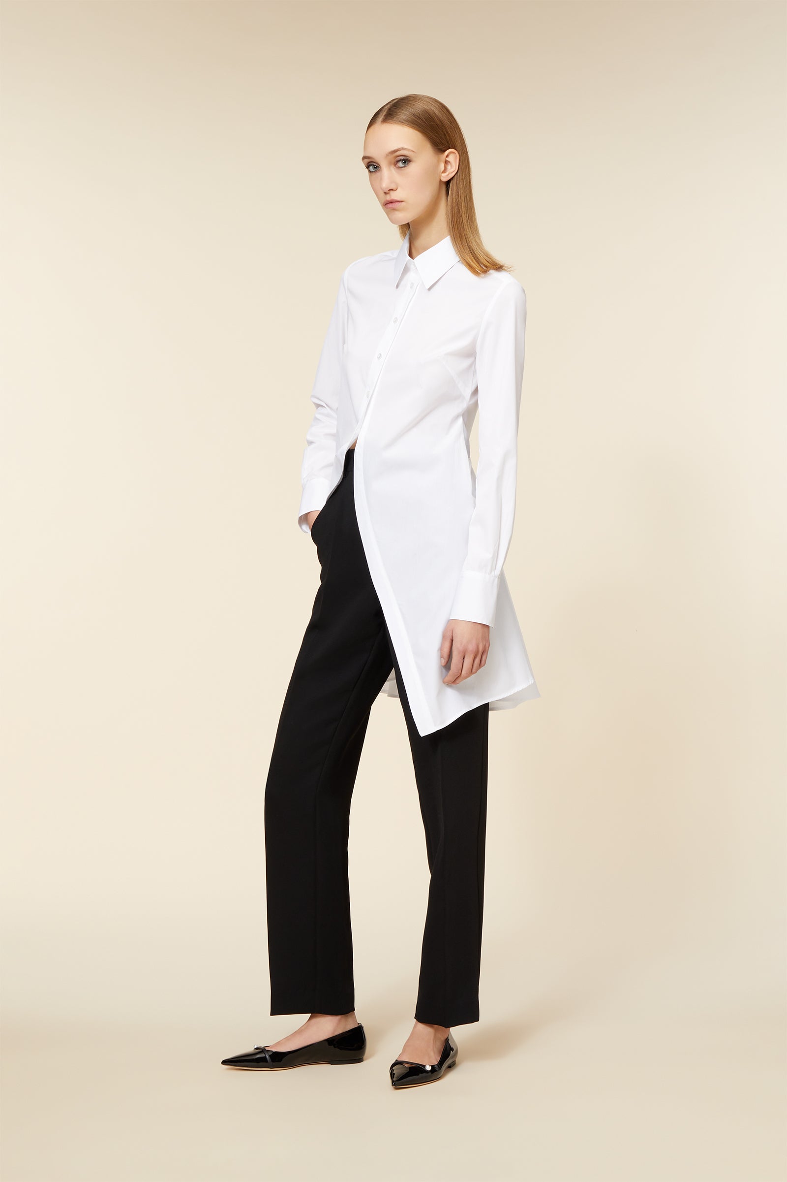 Long white button-up tunic paired with sleek black trousers and glossy black flats, set against a neutral background.