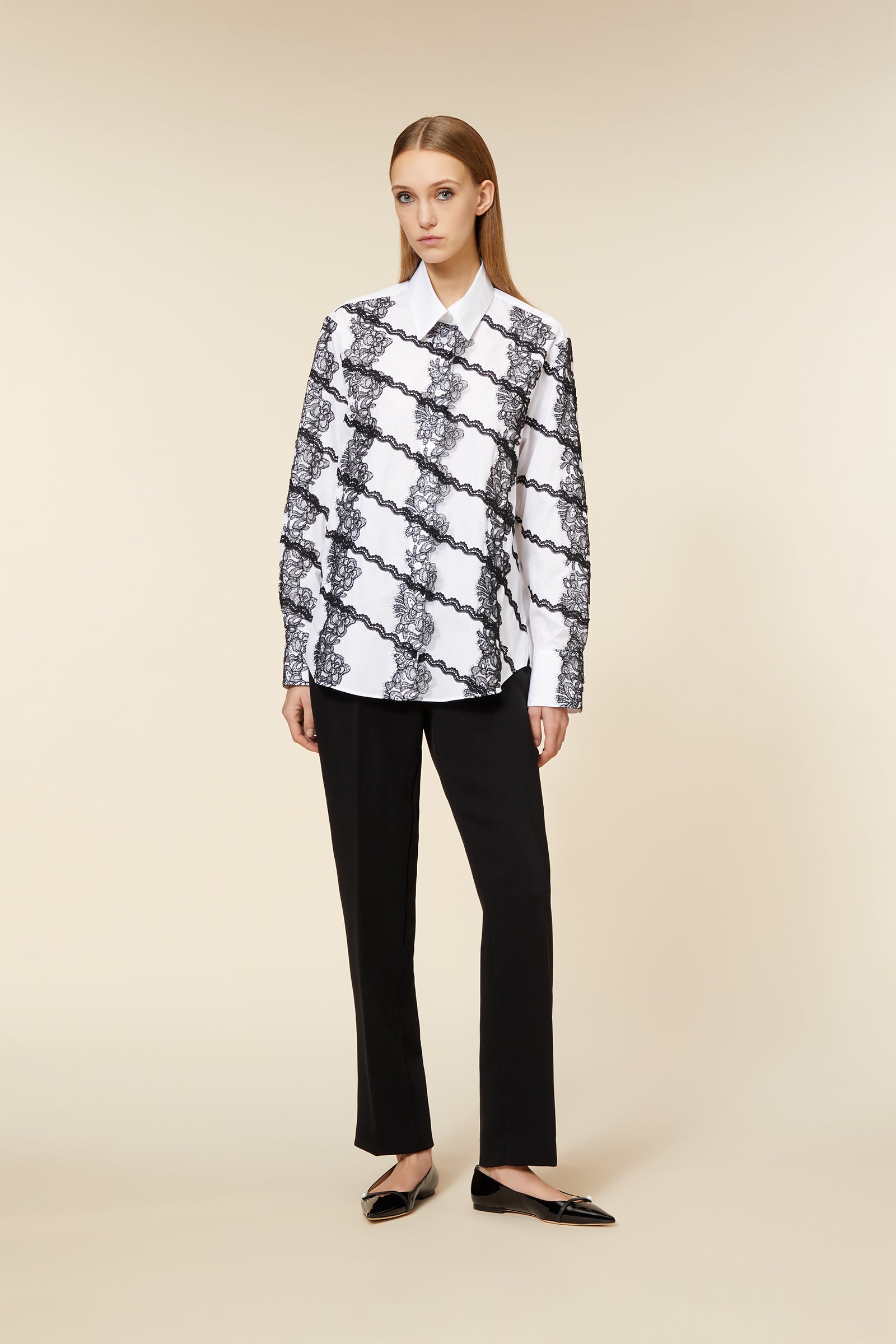 Model wearing a white shirt with intricate black lace pattern and classic black pants, styled with sleek, flat shoes.