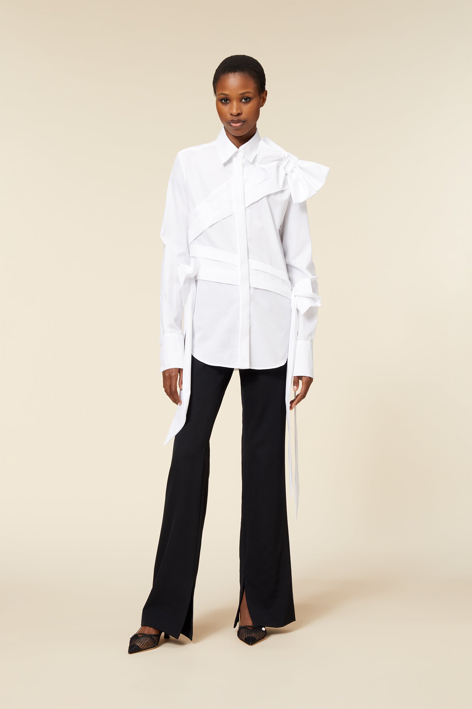 Model wearing a white avant-garde shirt with ribbon details and black wide-leg trousers, set against a neutral background.
