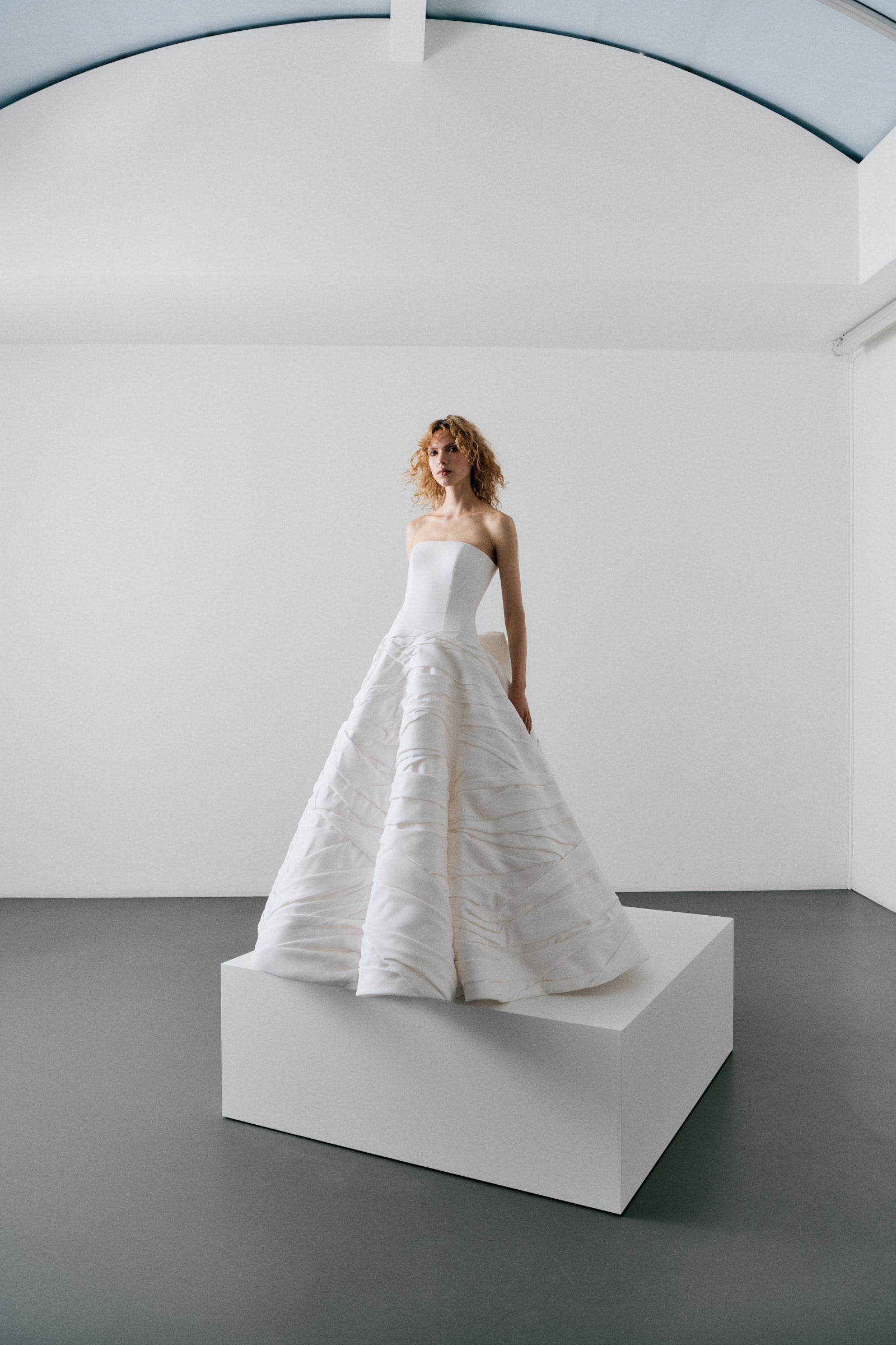 Woman in a white strapless gown standing on a white platform against a plain white wall.