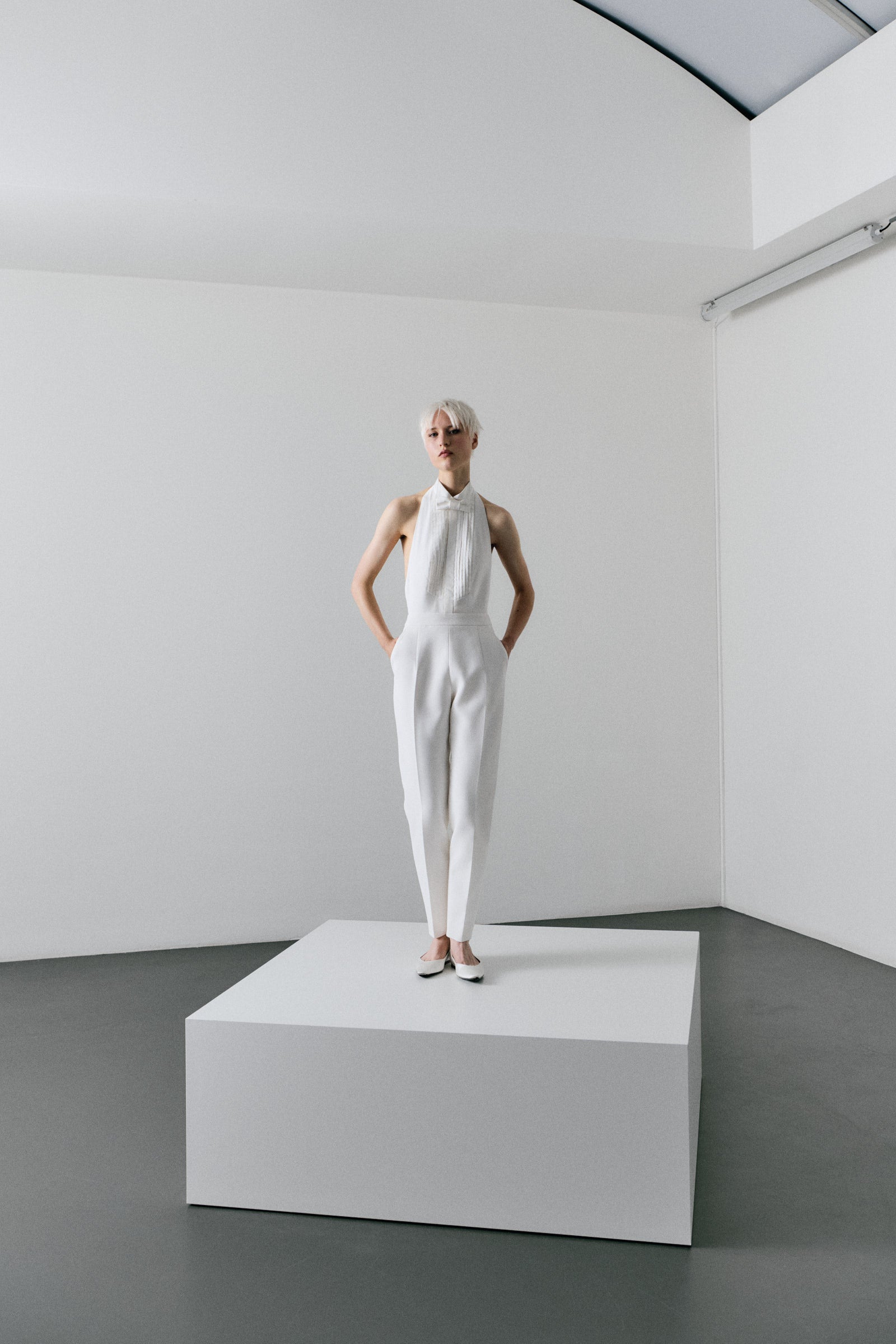 Person wearing a white outfit standing on a platform in a minimalistic room.