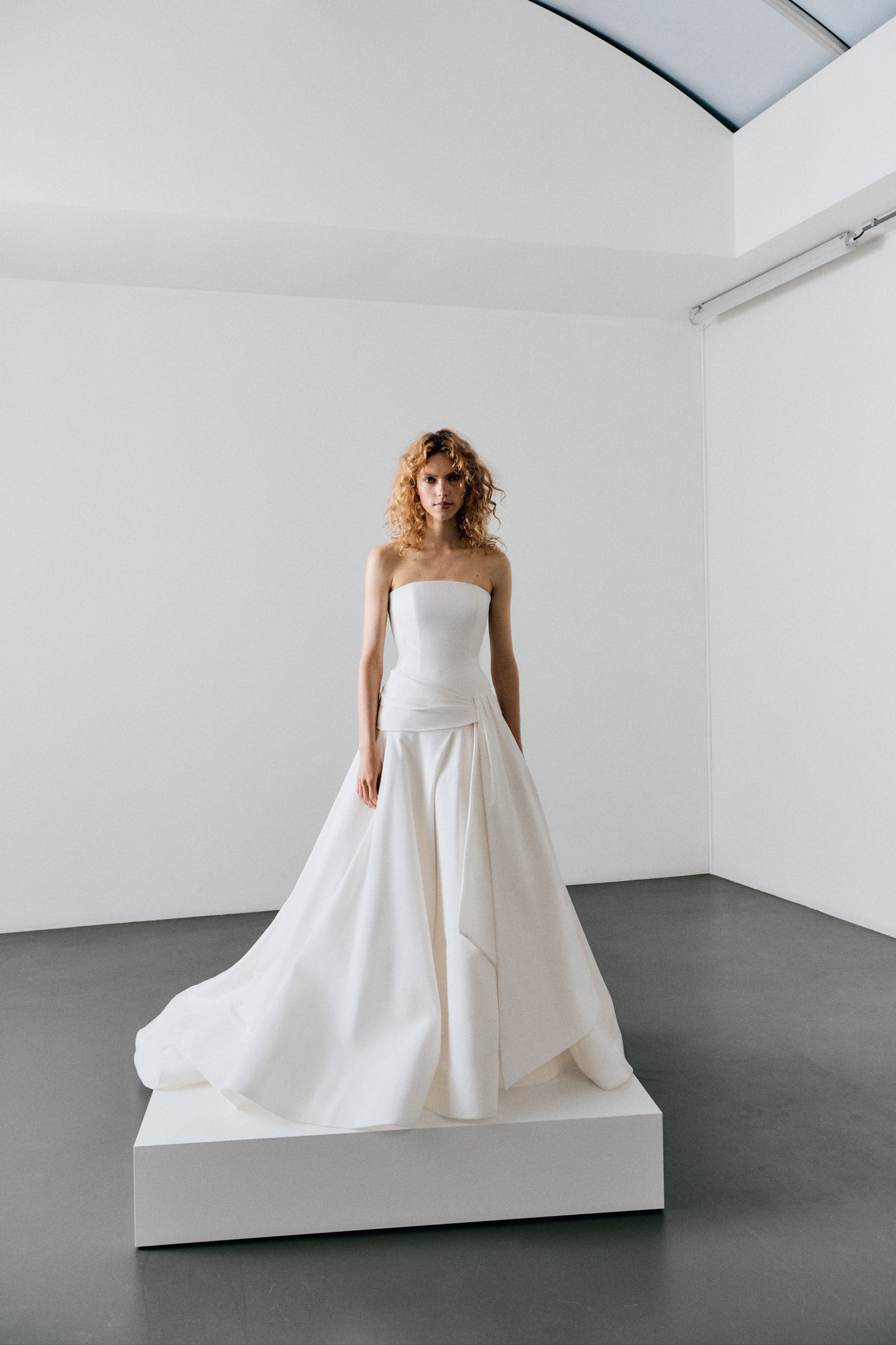 Woman in a white strapless dress standing on a platform in a minimalistic room.