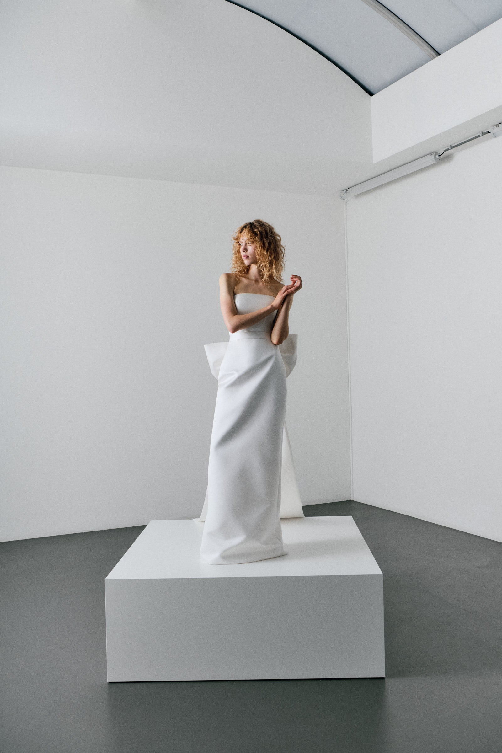 Woman in a white strapless dress standing on a platform in a minimalistic room.