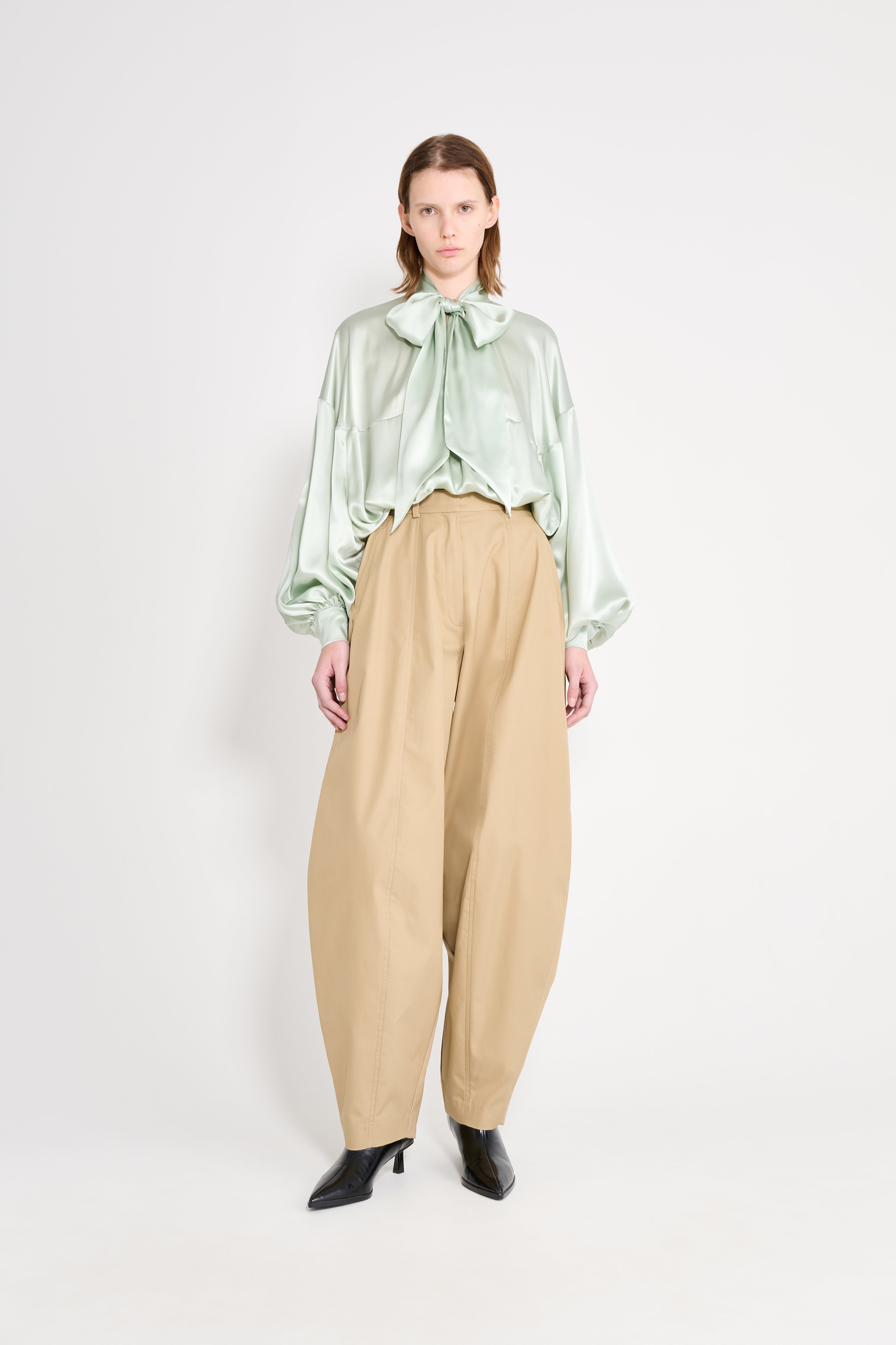Woman wearing a light green blouse with a bow and beige pants on a white background