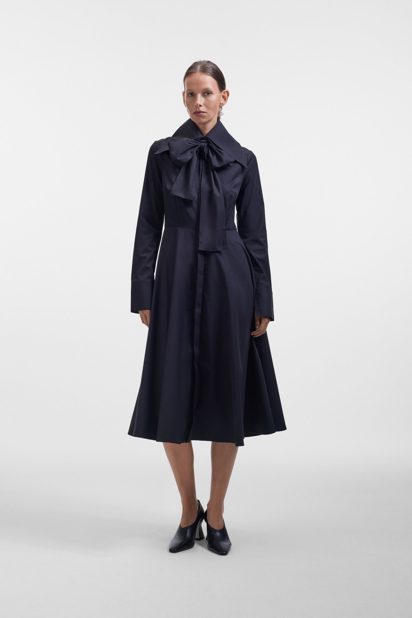 Model wearing a long black dress with a multi-layer collar and a bow detail on a white background.