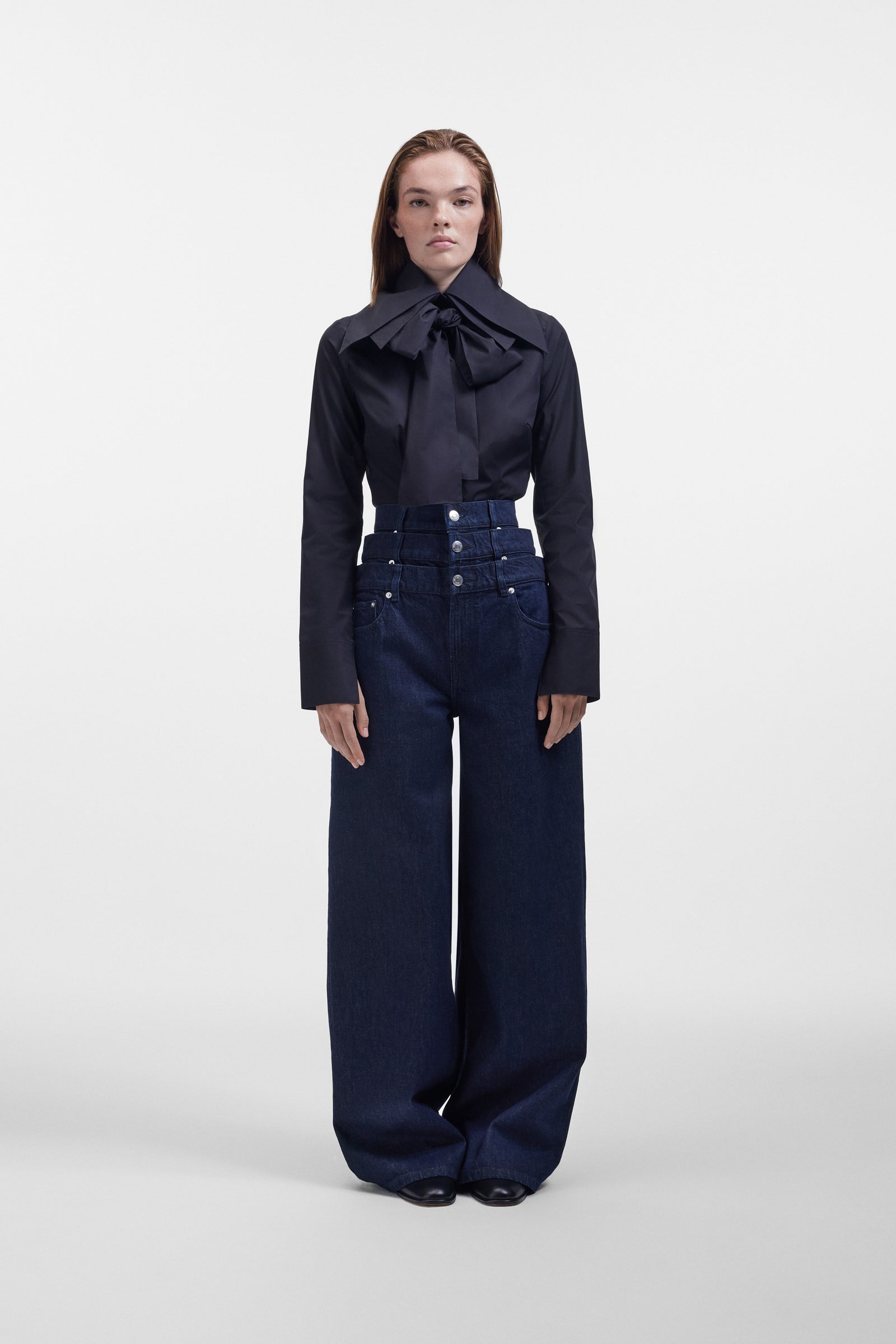 Model wearing a black long sleeve shirt with a bow on the collar, styled with dark denim pants on a white background.