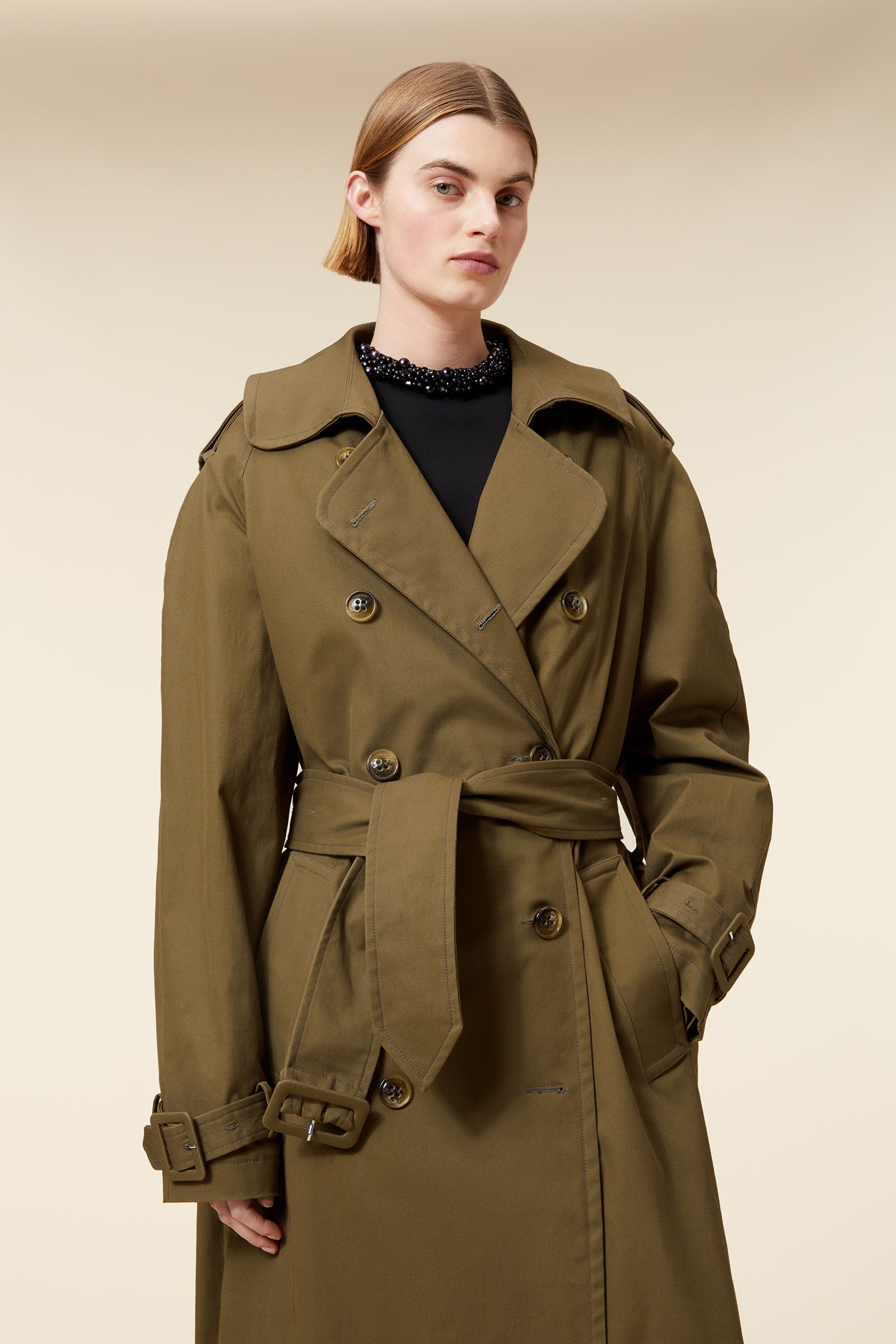 Olive green long trench coat featuring wide lapels, double-breasted buttons, and a belted waist, paired with black pants.