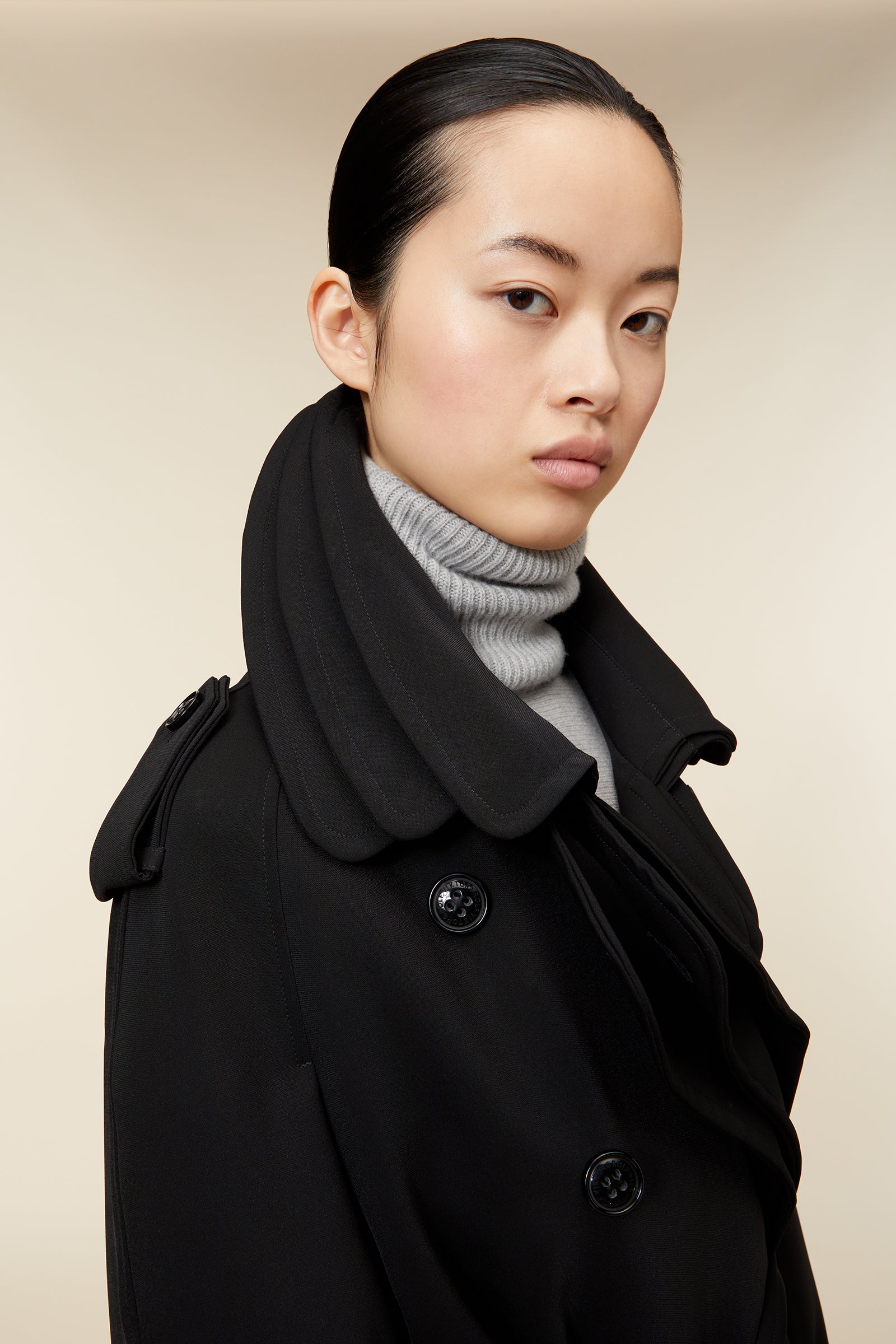 Long black trench coat with double-breasted buttons, belt, and notched lapel, worn over a gray turtleneck sweater.