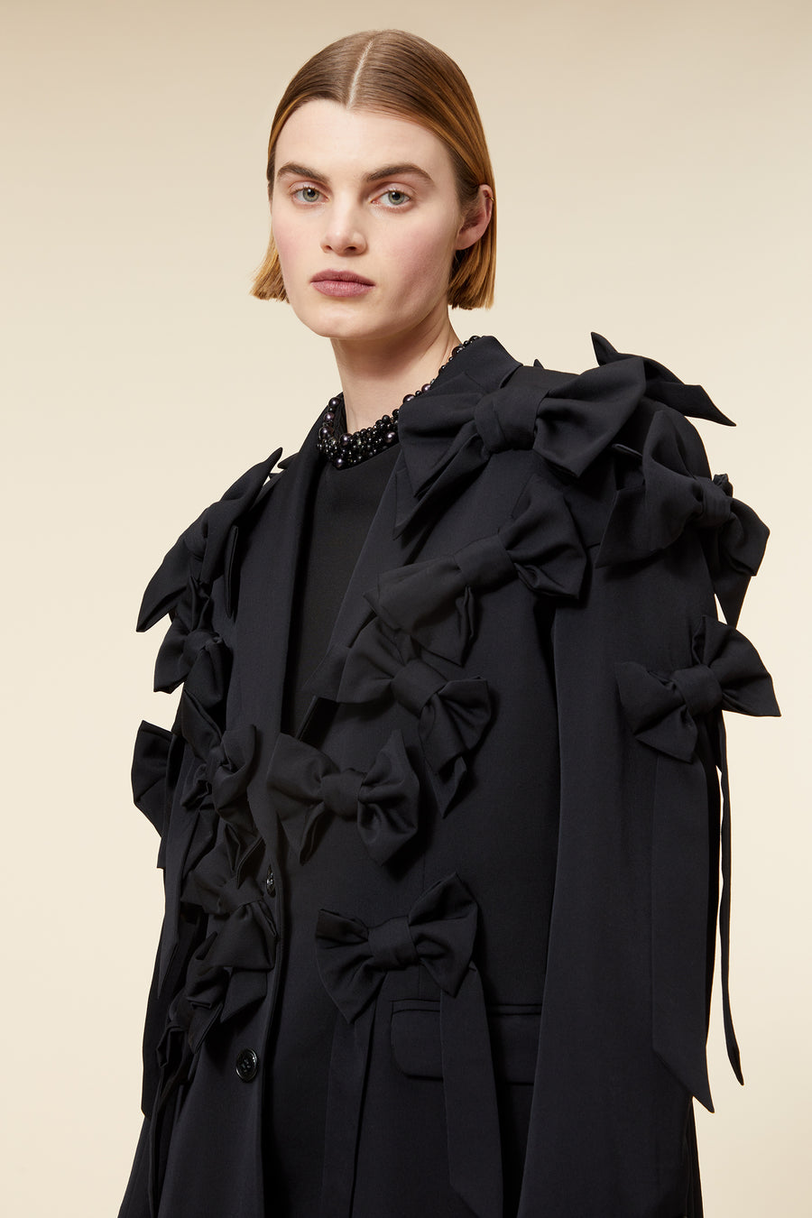 Elegant all-black suit with ruffled bow detailing, featuring tailored pants and paired with black loafers on beige background.
