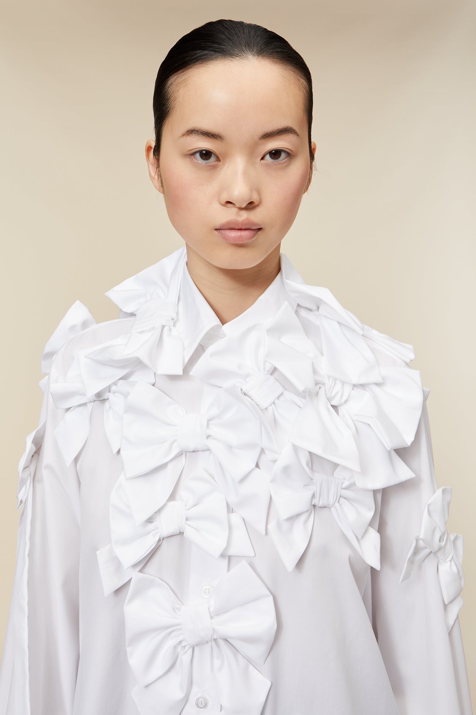 Model wearing a white ruffled blouse with bow details paired with black wide-leg trousers on a beige background.