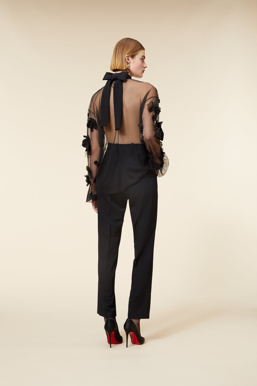 Model wearing a black sheer blouse with floral appliqué and matching trousers, posing against a neutral background.