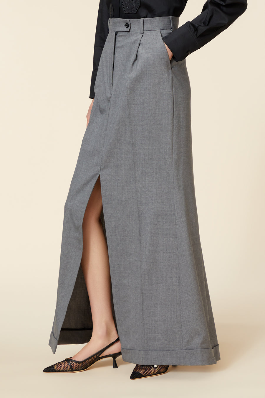 Gray wool high-waisted maxi skirt with front slit, tailored pleats, and black button detail, paired with mesh heels.