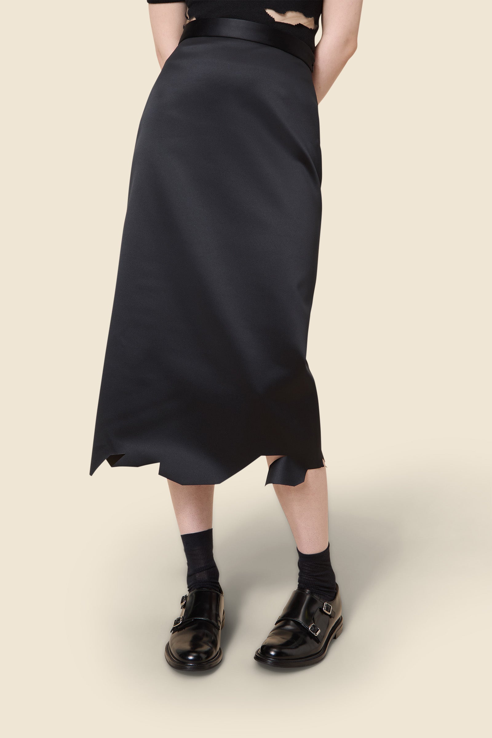 A sleek black A-line midi skirt with raw hem detailing, paired with shiny black monk strap shoes and black socks.