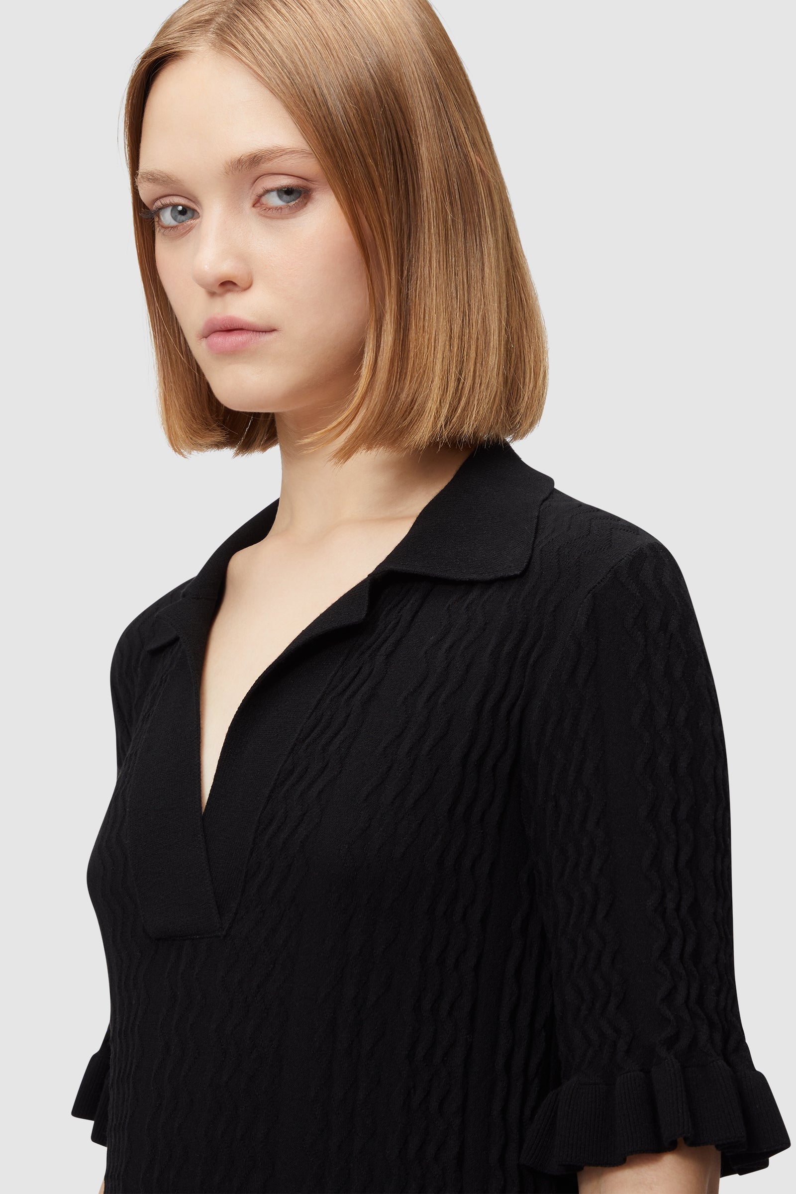 Woman wearing a textured black dress with a collared neckline, wavy pattern, and ruffled sleeves, light background.