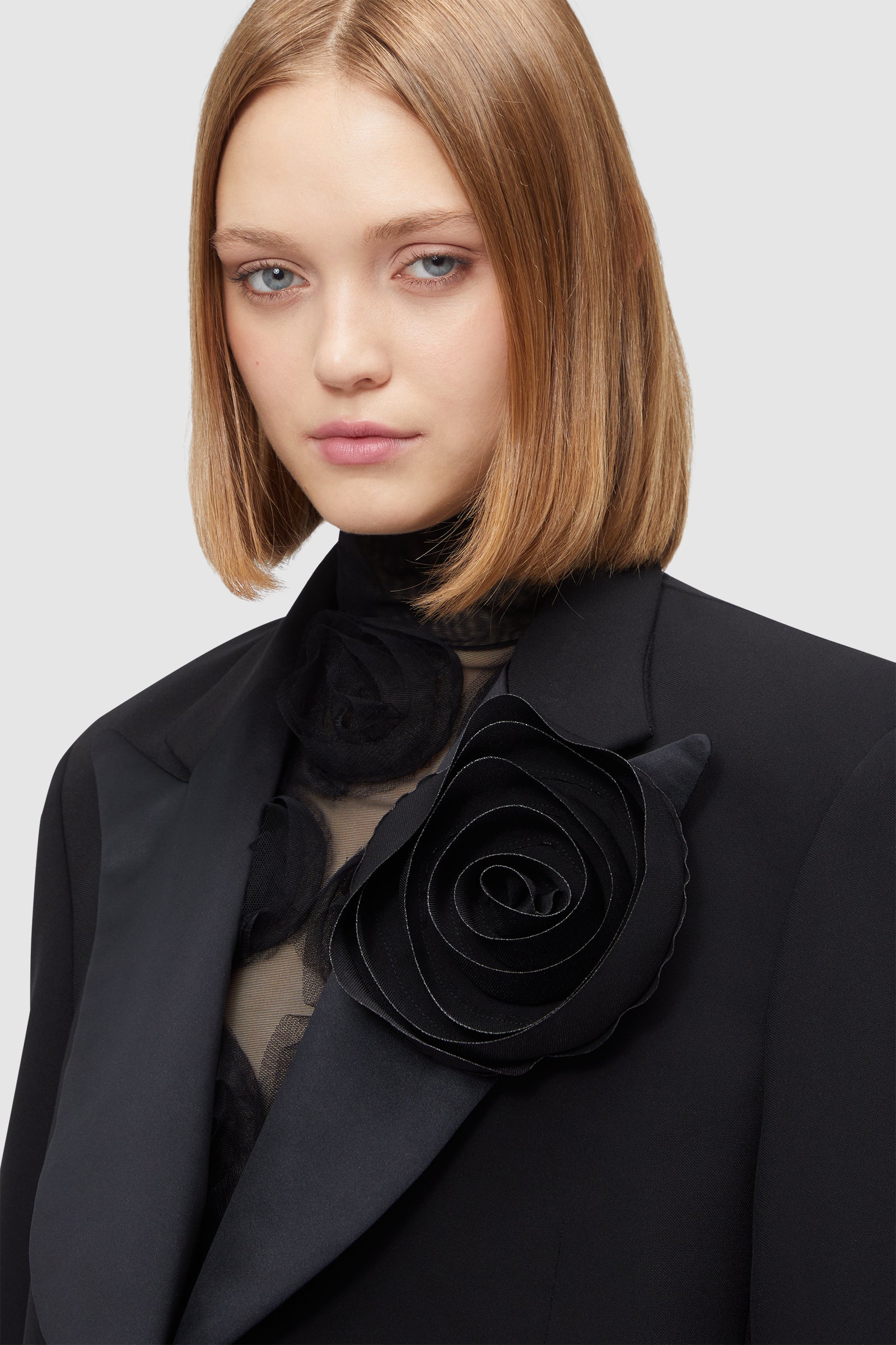 Black blazer features an oversized satin floral brooch and sheer blouse, adding elegance to its structured design.