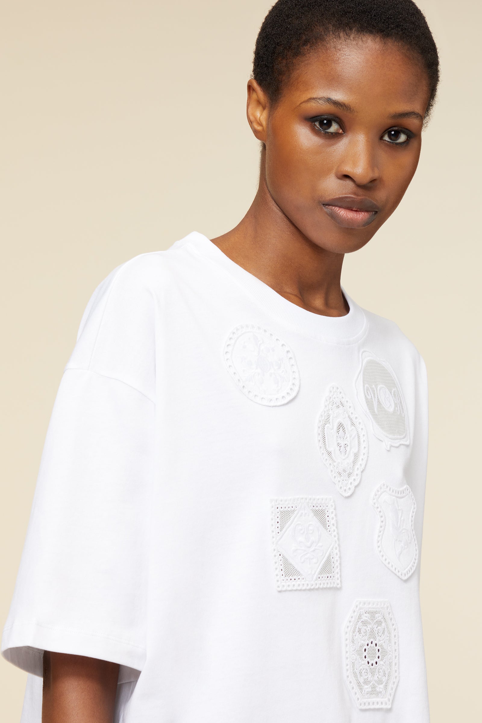 White cotton t-shirt with intricate lace appliqué motifs, featuring a relaxed fit and crew neckline, against a beige backdrop.