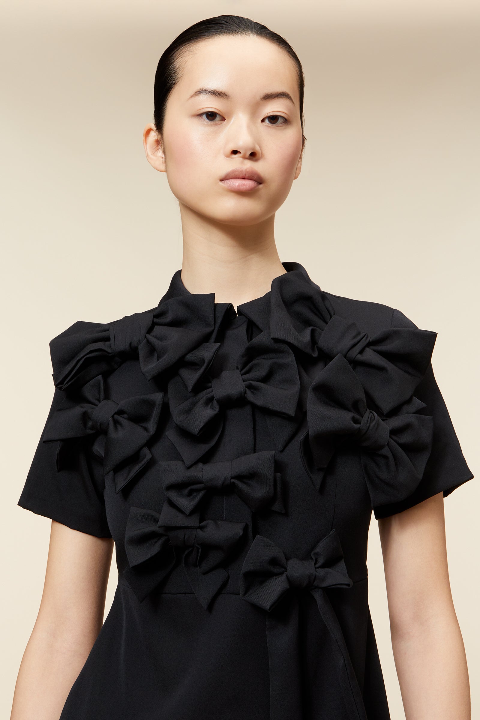 Model wearing elegant short black dress with large fabric bows, set against a neutral background.