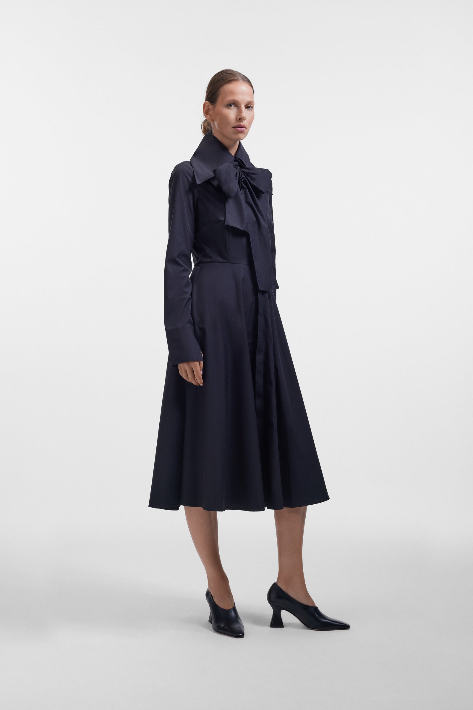 Model wearing a long black dress with a multi-layer collar and a bow detail on a white background.