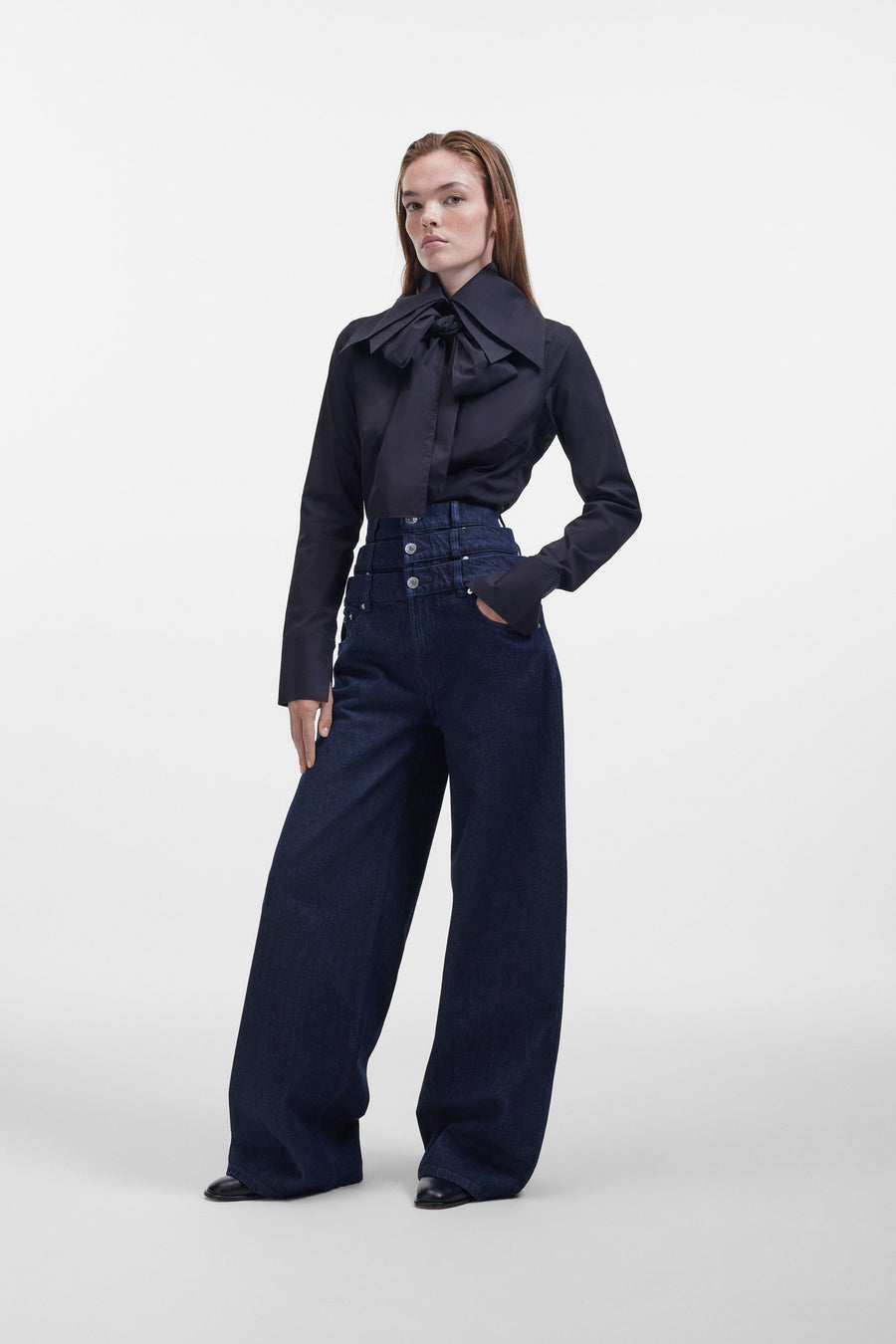 Model wearing a black long sleeve shirt with a bow on the collar, styled with dark denim pants on a white background.