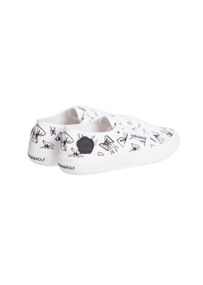 White canvas sneakers adorned with black bow illustrations, featuring branding details on the heel and a rubber sole.