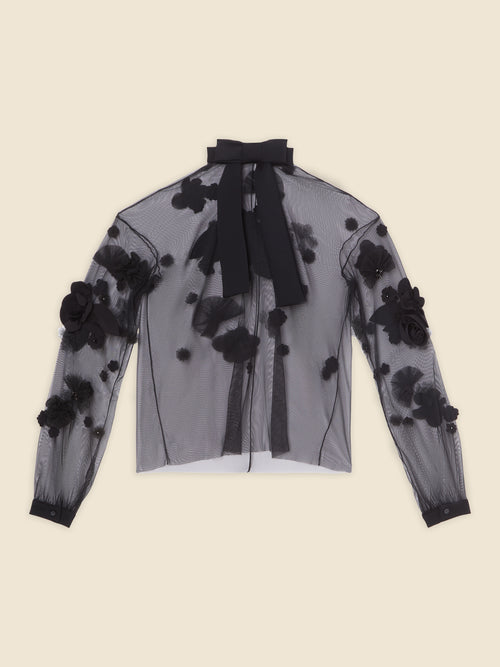 Sheer black blouse with 3D floral appliqué design and bow tie collar on creamy beige background.