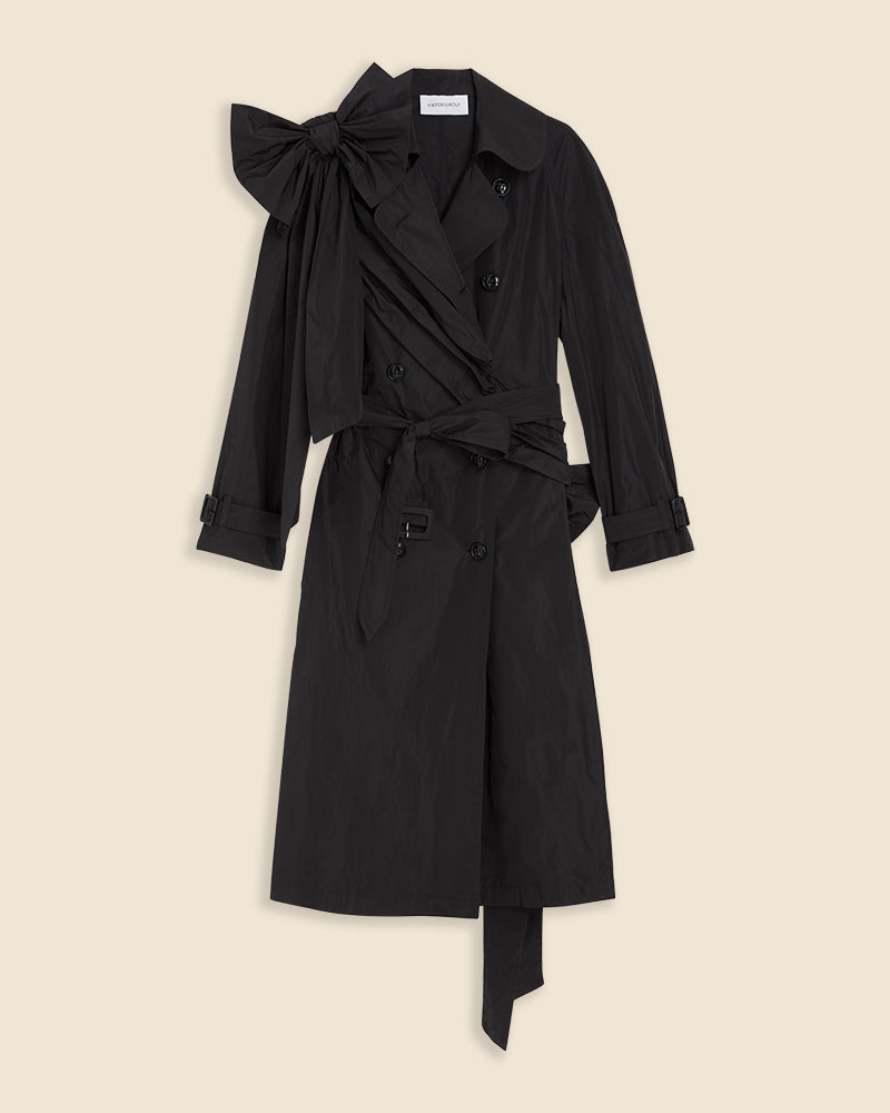 Black trench coat with oversized bow detailing on the shoulder and double-breasted button design, featuring a classic waist belt.