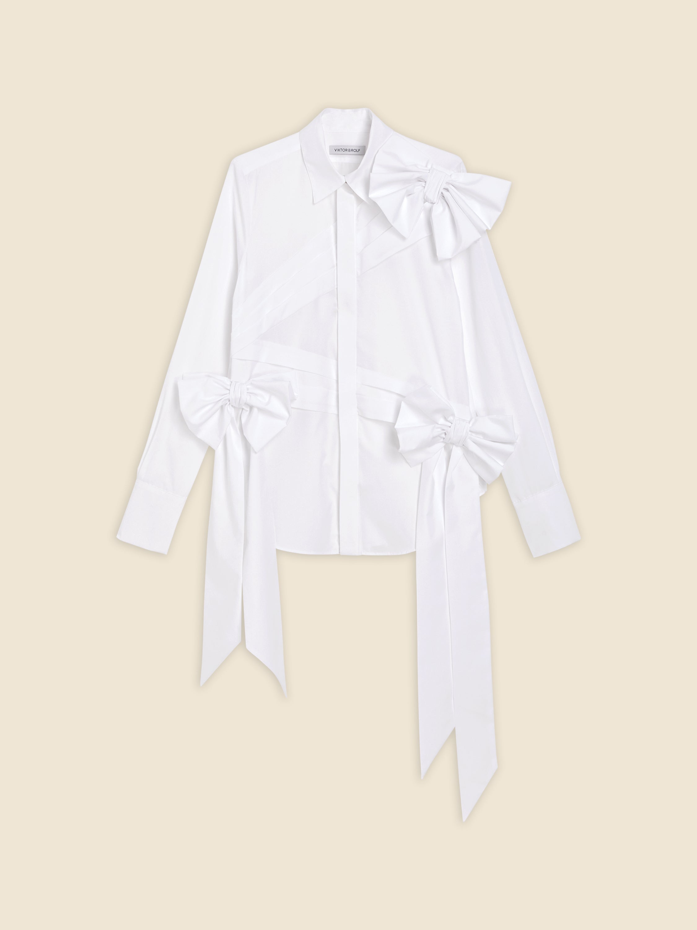 White long-sleeve shirt with decorative bows, featuring buttoned front and classic collar on beige background.