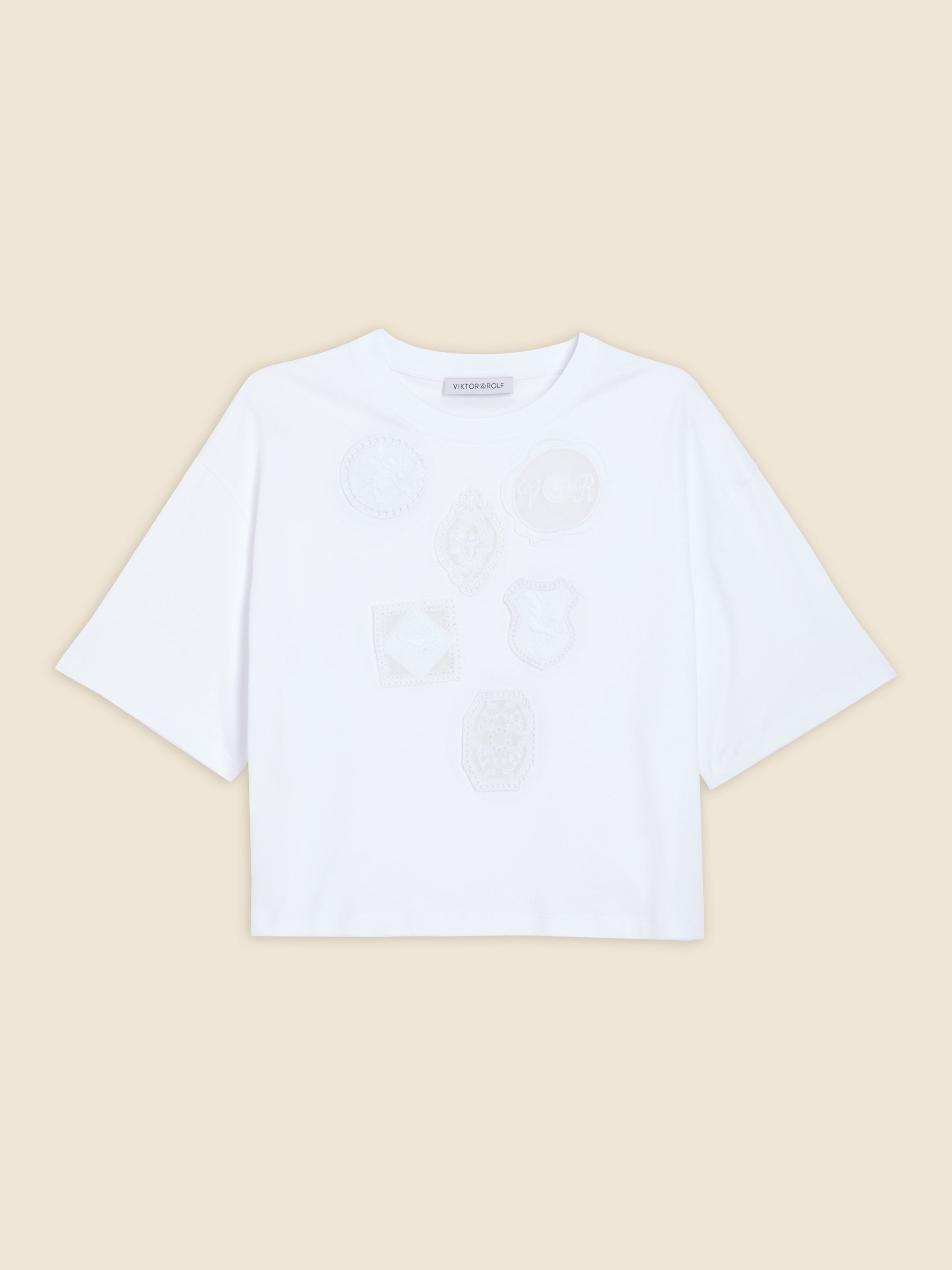 White shirt featuring subtle embroidered badge motifs on the front, set against a plain beige background, short-sleeved style.