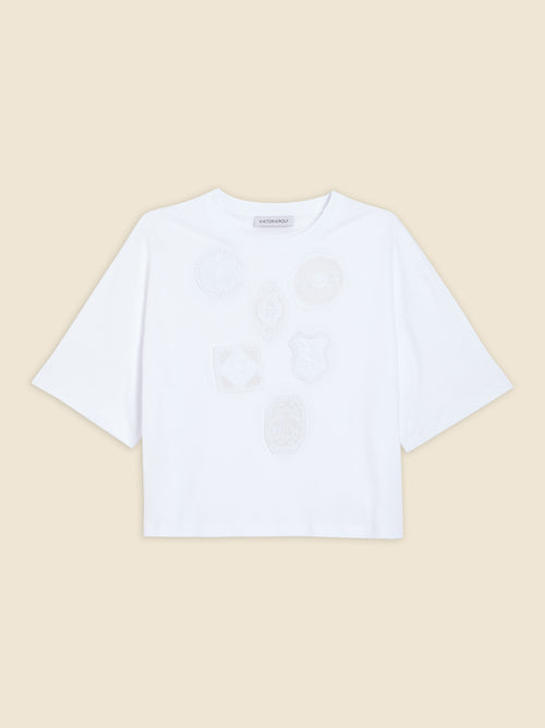 White shirt featuring subtle embroidered badge motifs on the front, set against a plain beige background, short-sleeved style.