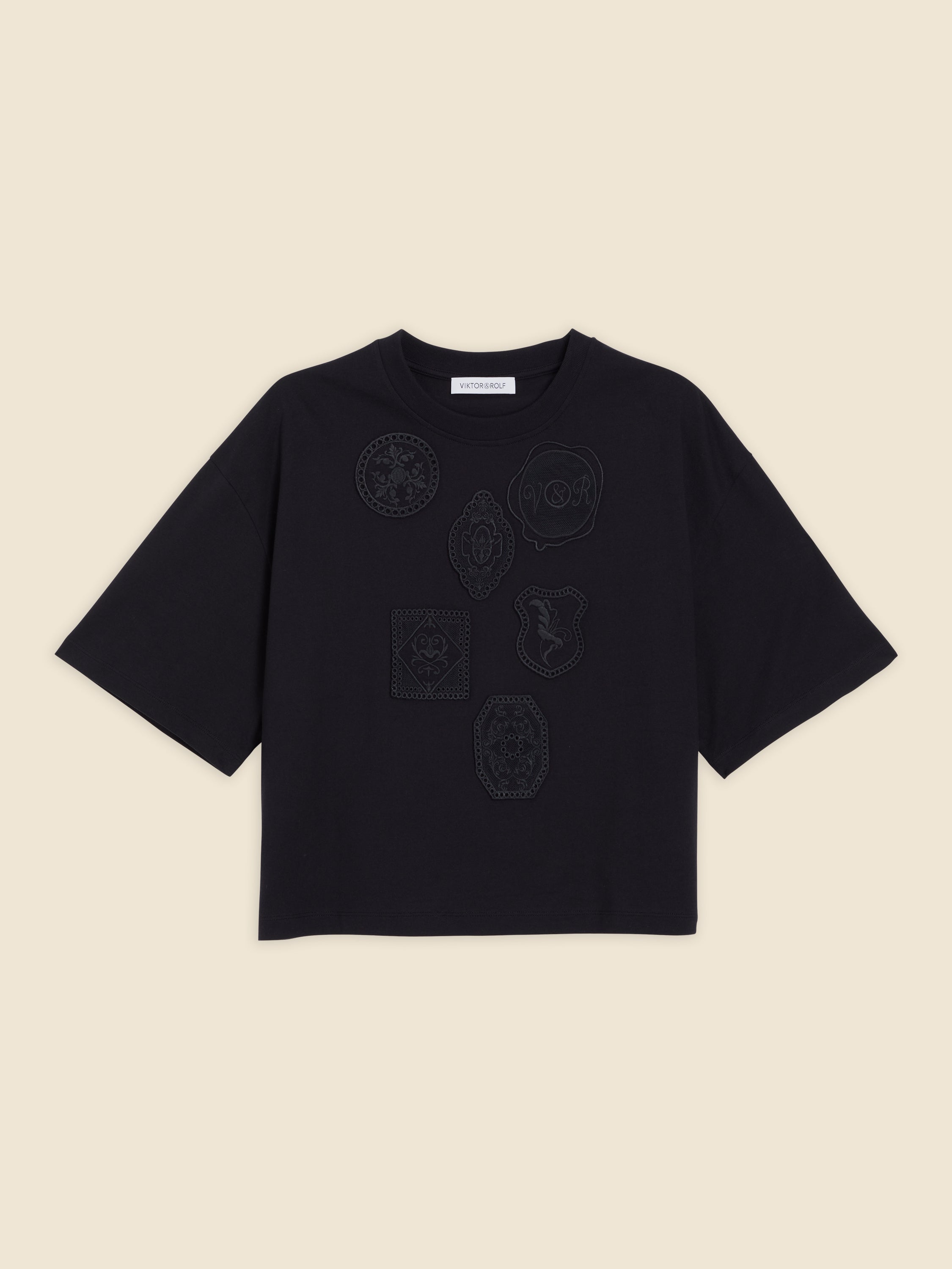 Black cotton T-shirt featuring intricate embroidered patches with minimalist designs against a plain beige background.