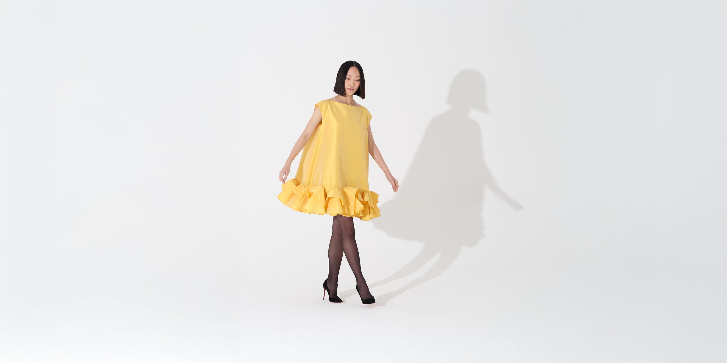 Model wearing a yellow boxy dress with ruffles at the bottom on a white background.