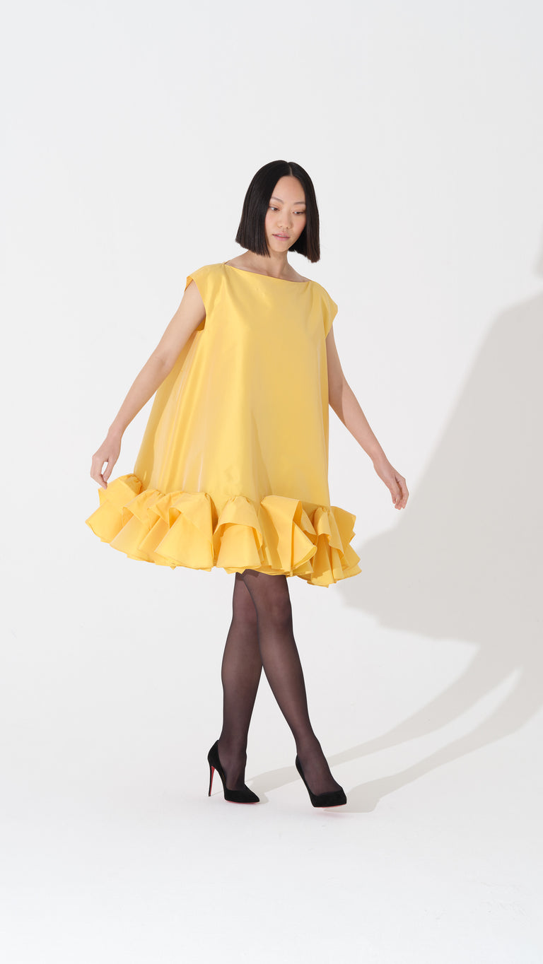 Model wearing a yellow boxy dress with ruffles at the bottom on a white background.