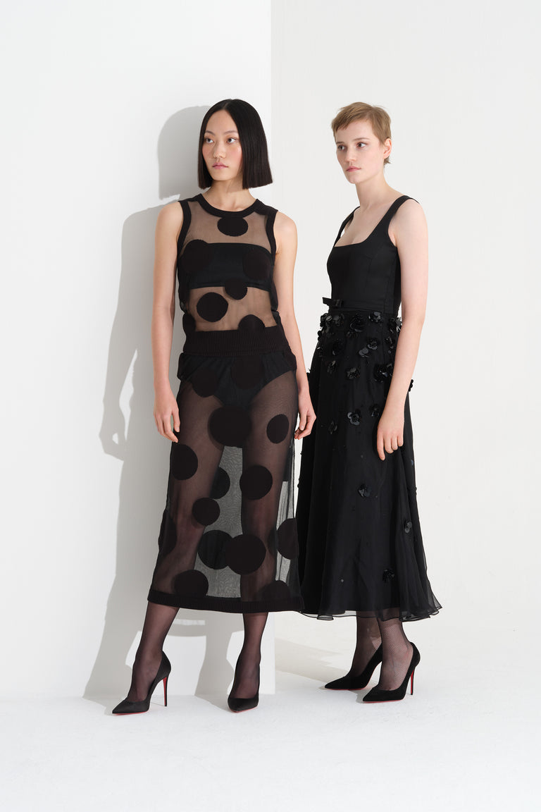 Two models wearing a black sheer polka dot dress and a long black night dress, standing on a white background.