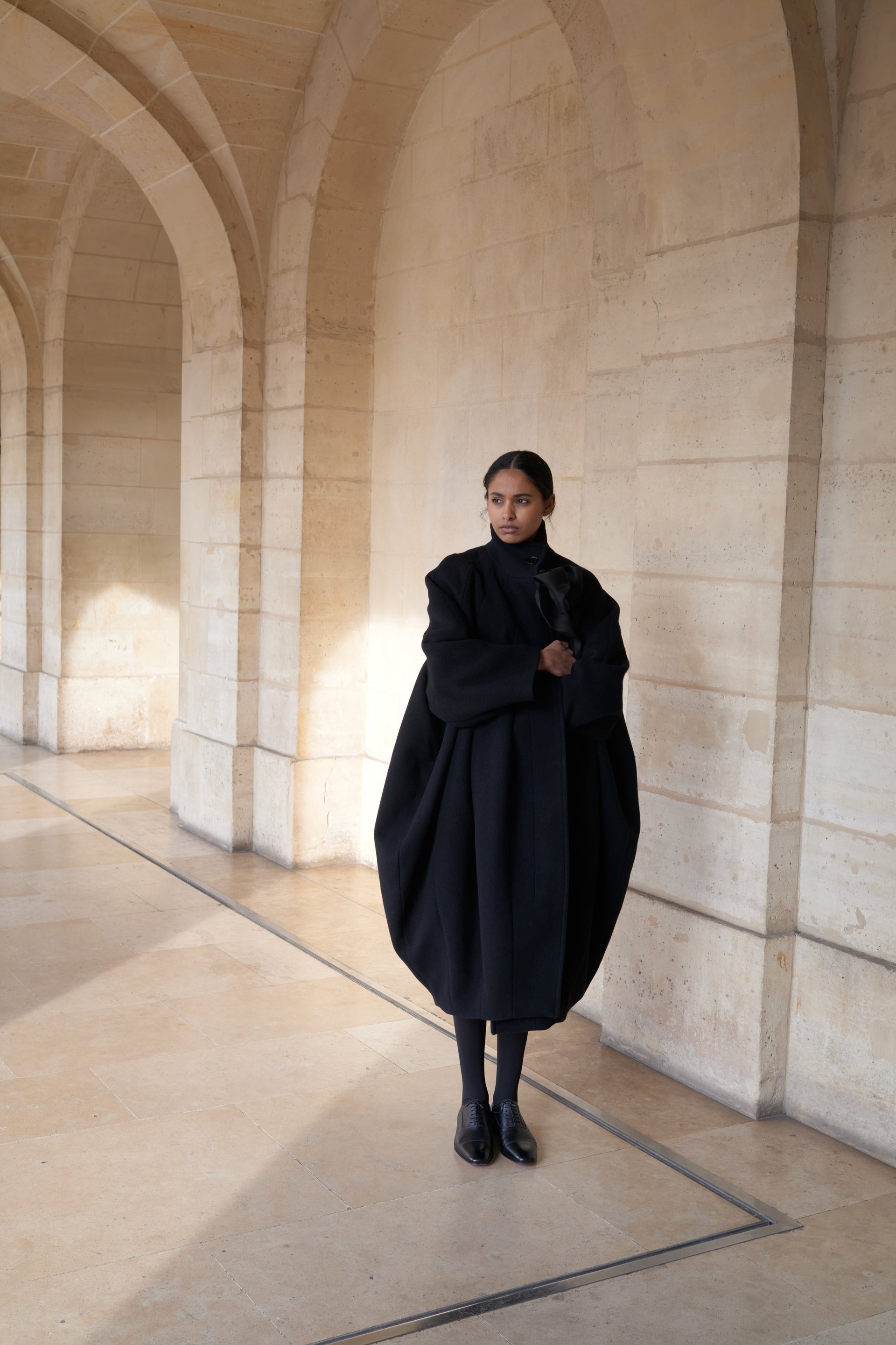 Model wearing a black oversized coat in front of a beige wall.