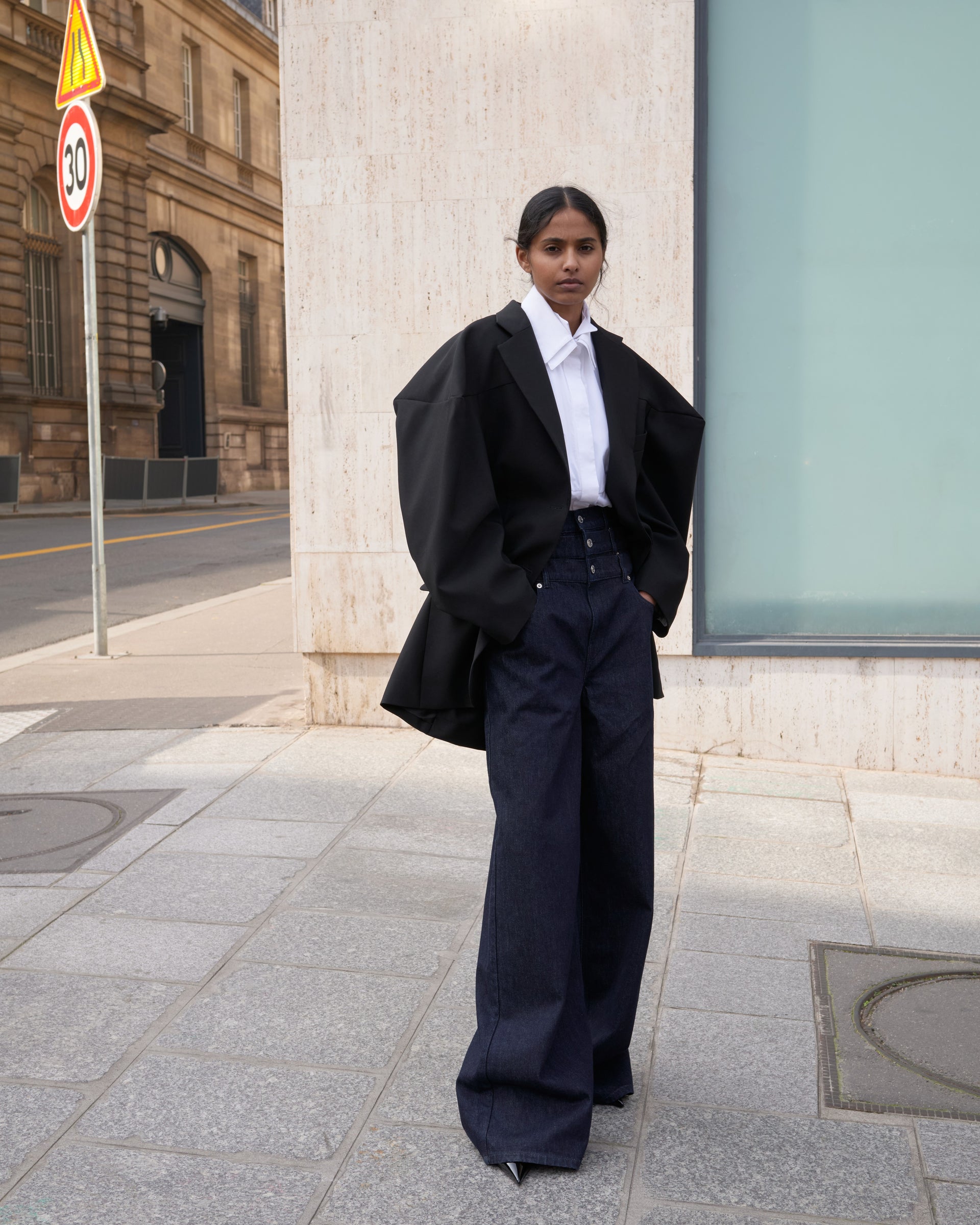 Person wearing a black blazer and navy pants standing on a city street.