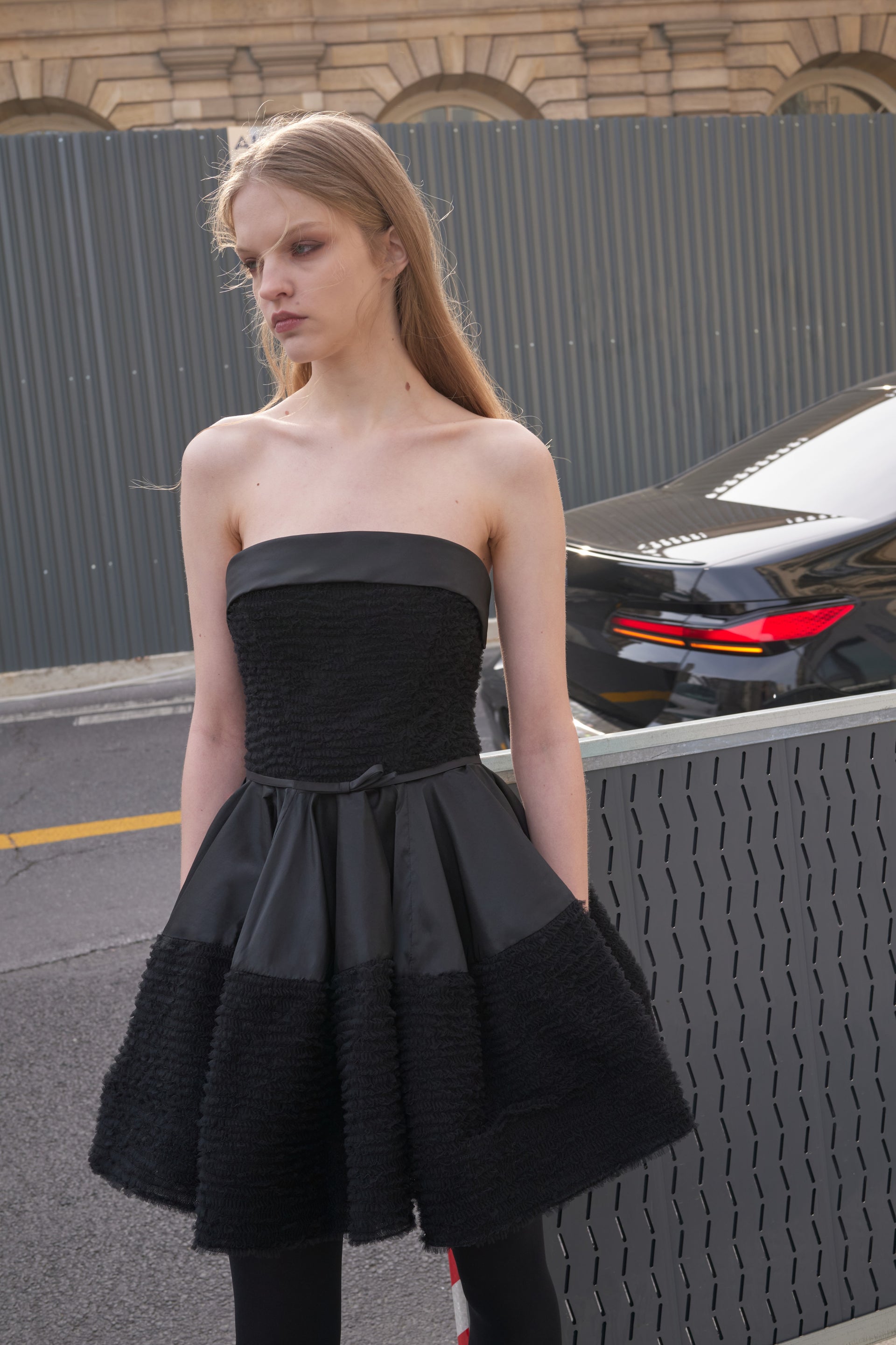 Woman wearing a black strapless dress standing on a street with a car in the background.