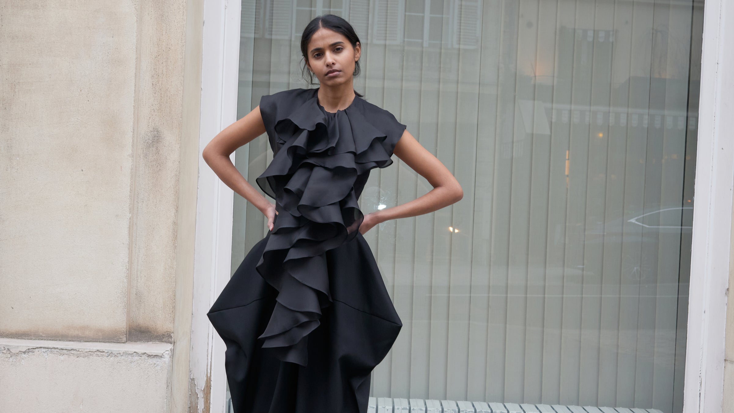 Woman wearing a black ruffled dress standing against a building exterior.