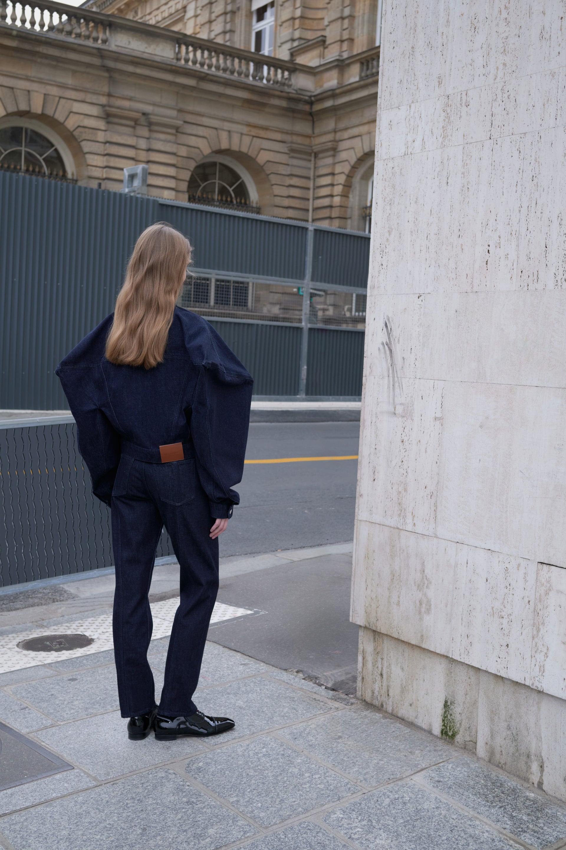Person in a navy denim suit walking away from a building with a fence.