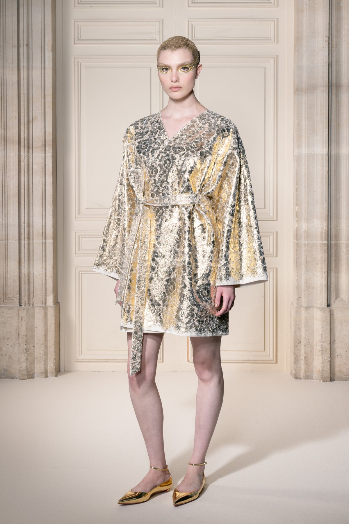 Una modella indossa un abito corto in stile kimono, con cintura, ricoperto di paillettes metalliche dorate, argentate e beige, in piedi davanti a una parete rivestita di pannelli in stile classico.