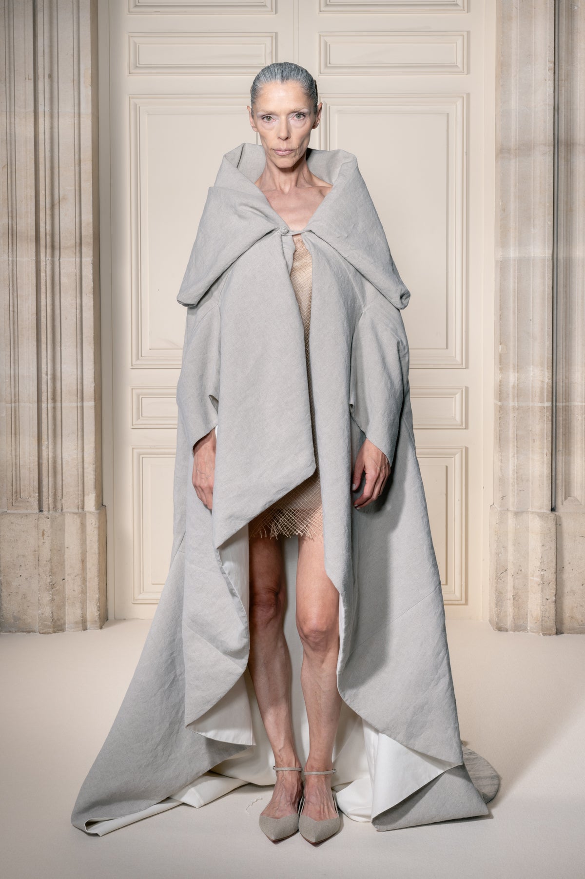 Una modella posa in un interno sontuoso con pareti rivestite in legno color crema, indossando un voluminoso scialle o mantello drappeggiato grigio chiaro sopra un abito color carne, abbinato a scarpe grigie a punta.