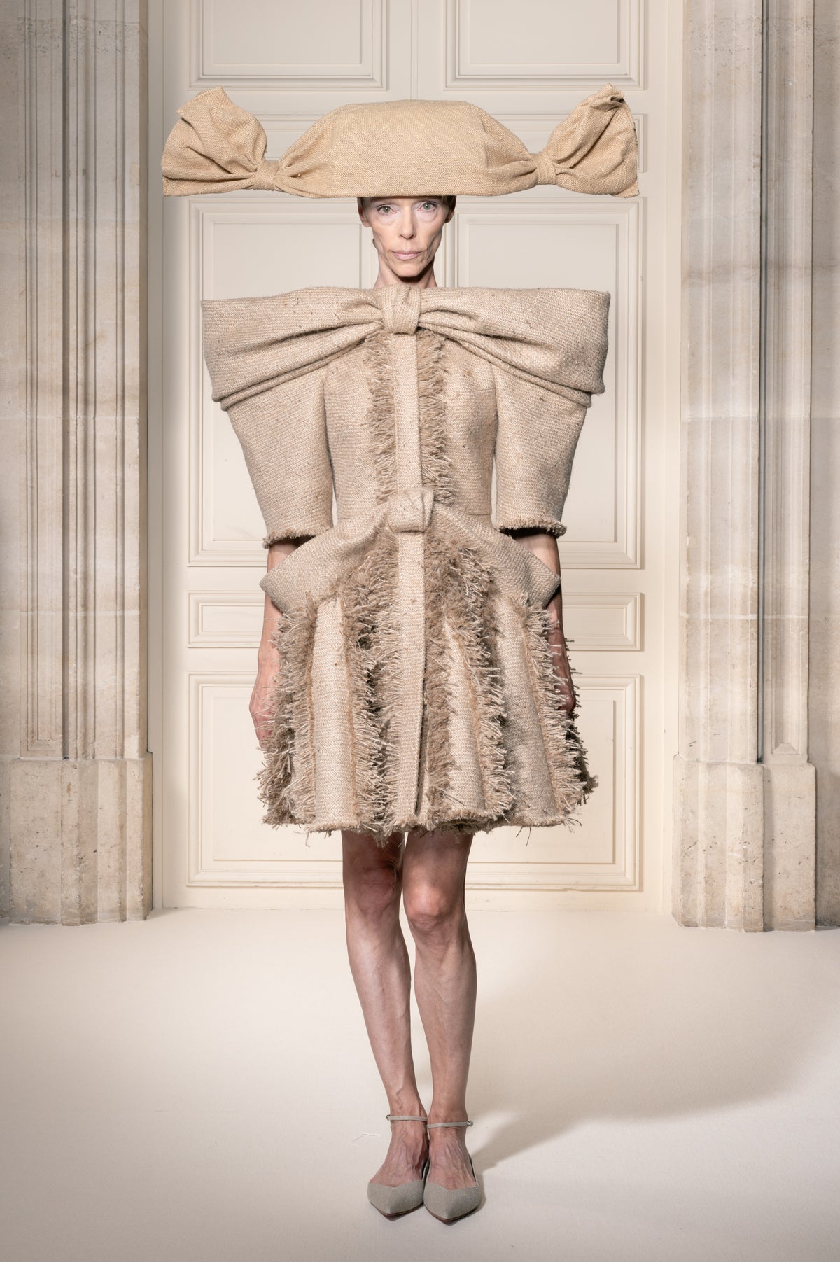 In un sontuoso interno con colonne classiche, una modella indossa un cappotto beige scultoreo con dettagli a fiocco oversize, un cappello a tesa larga abbinato e scarpe grigie a punta.