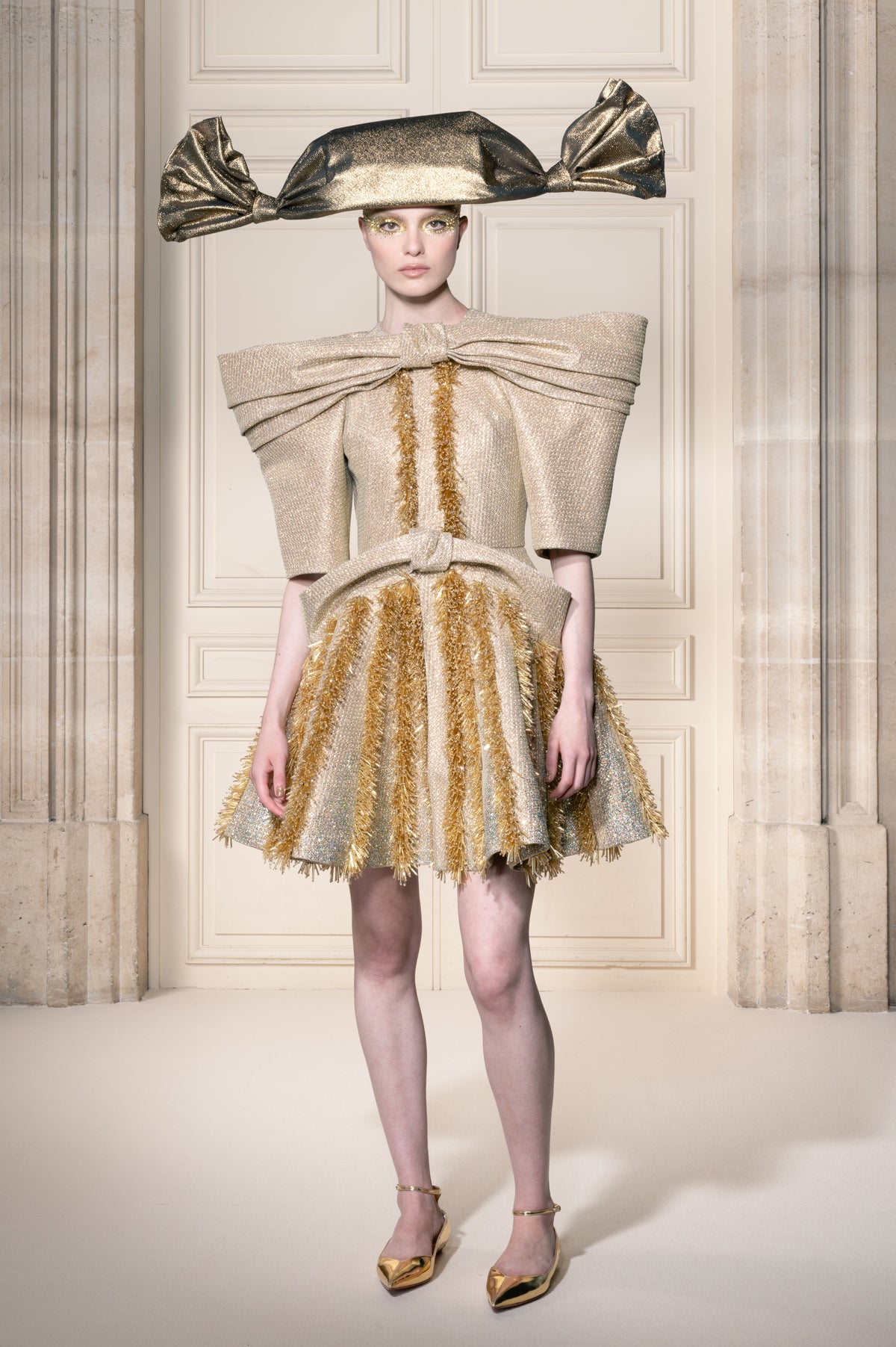 Una modella posa in un sontuoso interno con colonne classiche, indossando un miniabito beige e oro dalla struttura molto definita con pieghe vistose sulle spalle, un cappello a tesa larga abbinato e scarpe a punta dorate.