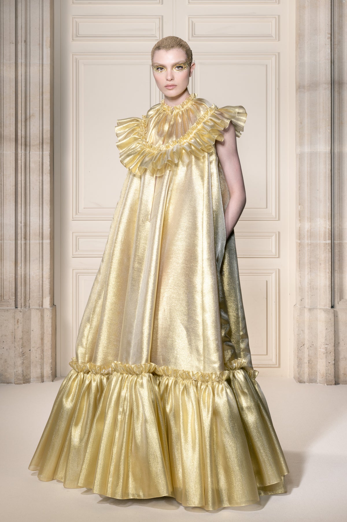Una modella indossa un voluminoso abito color oro metallizzato con elaborate balze, in piedi davanti a una parete rivestita di pannelli in stile classico.