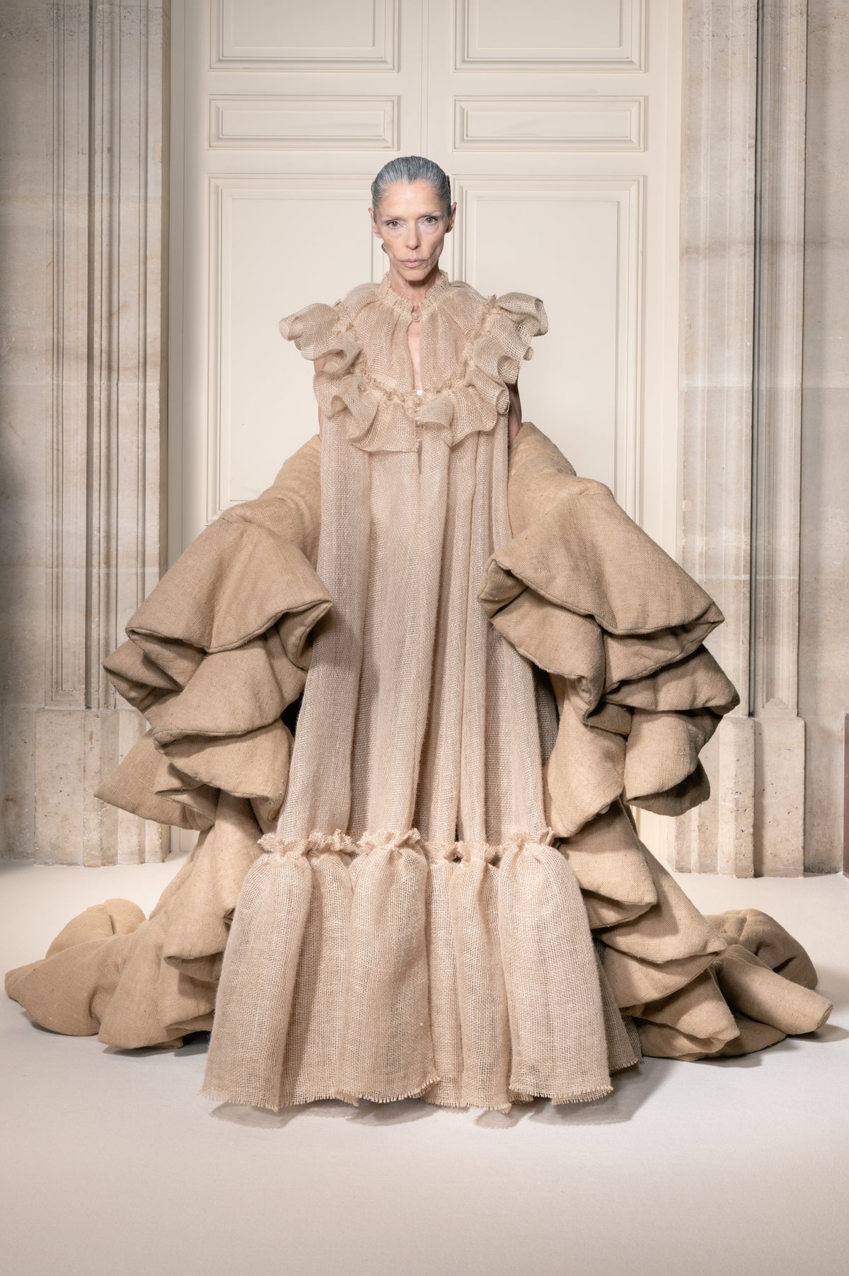 Una modella posa in una grande sala indossando un abito beige oversize a balze, caratterizzato da un elaborato strato di tessuto e un alto colletto a balze.