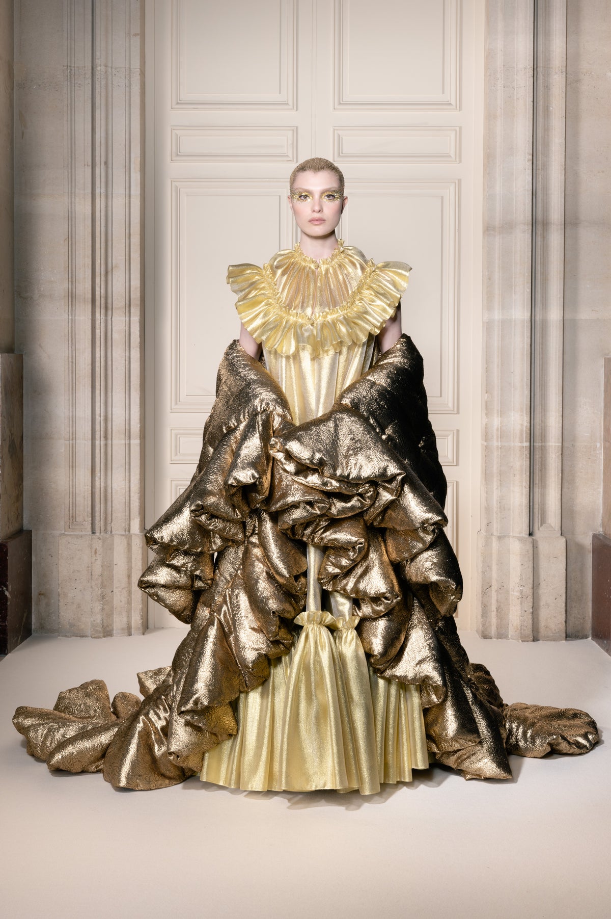 Una modella posa in una grande sala indossando un elaborato abito color oro metallizzato con sontuose balze a strati e un voluminoso drappeggio sovrapposto, fotografata in un interno simmetrico con colonne in pietra.