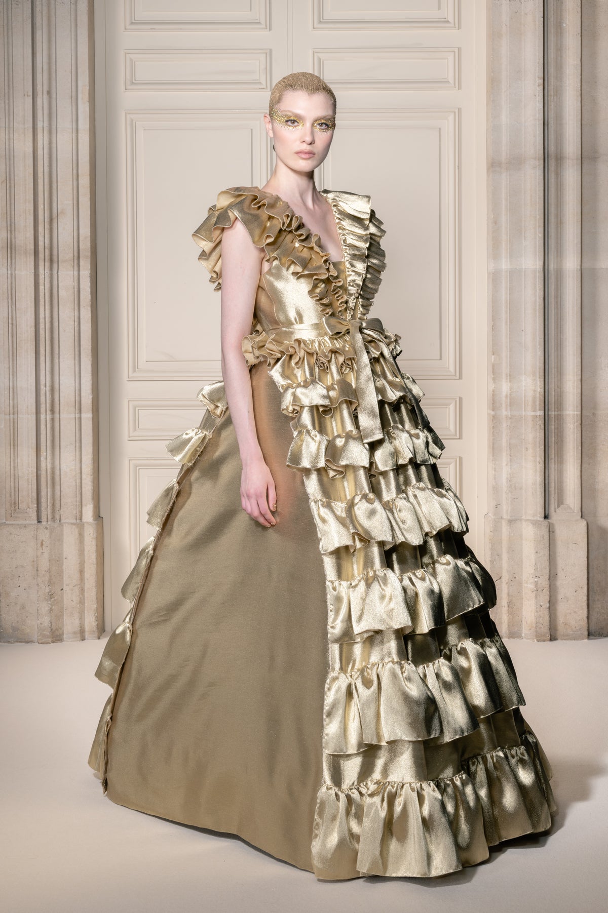 Una modella indossa un sontuoso abito lungo fino ai piedi color oro, con un corpetto strutturato e a balze e una gonna a cascata a strati, in posa davanti a una parete rivestita in legno in stile classico.