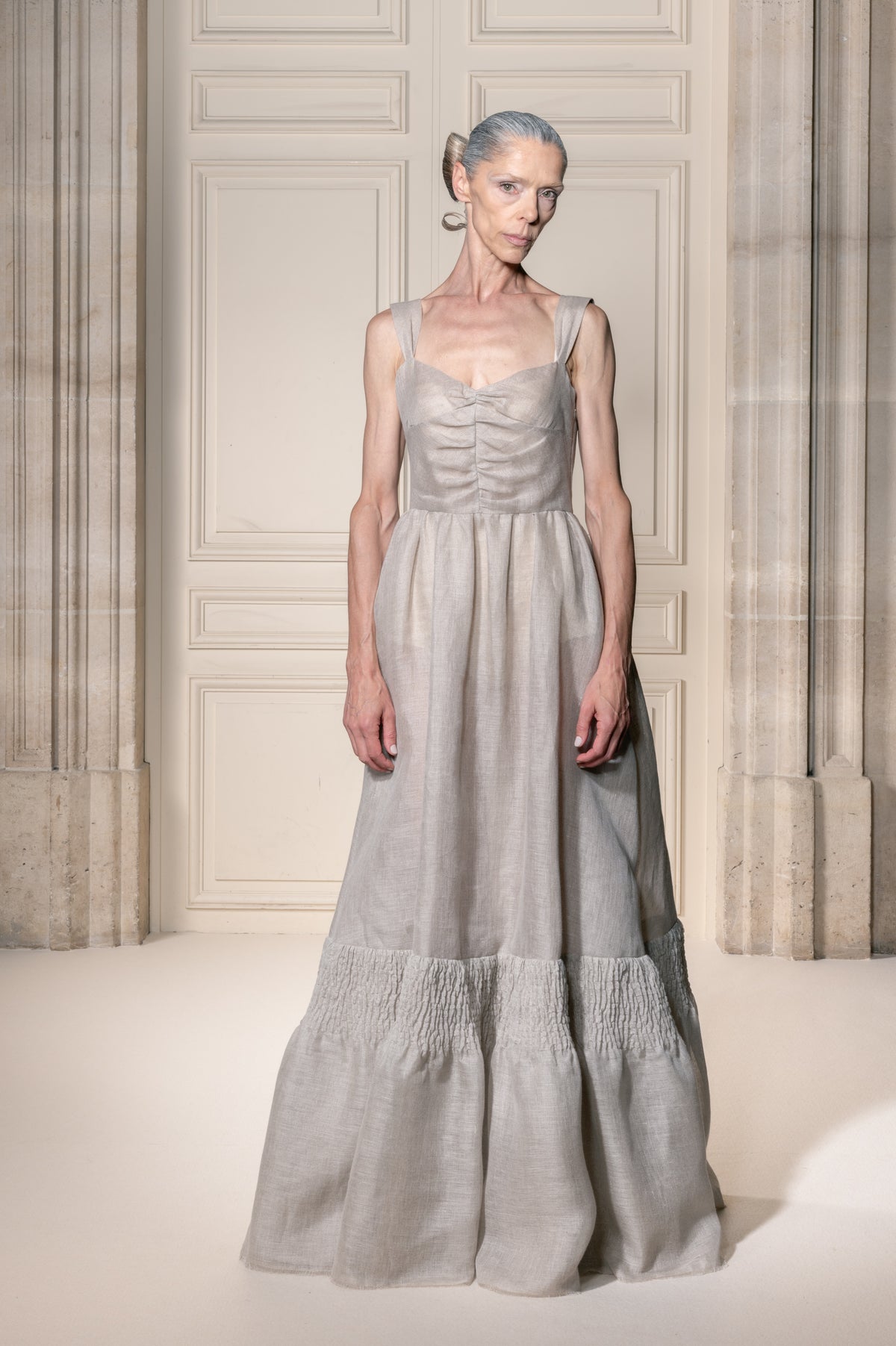 Una modella indossa un abito lungo di lino grigio chiaro con corpetto arricciato e gonna a balze, in piedi davanti a una classica porta a pannelli color crema.