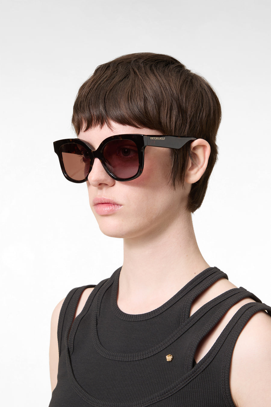 Short dark hair woman wearing oversized black sunglasses and a sleeveless ribbed black top against a beige background.
