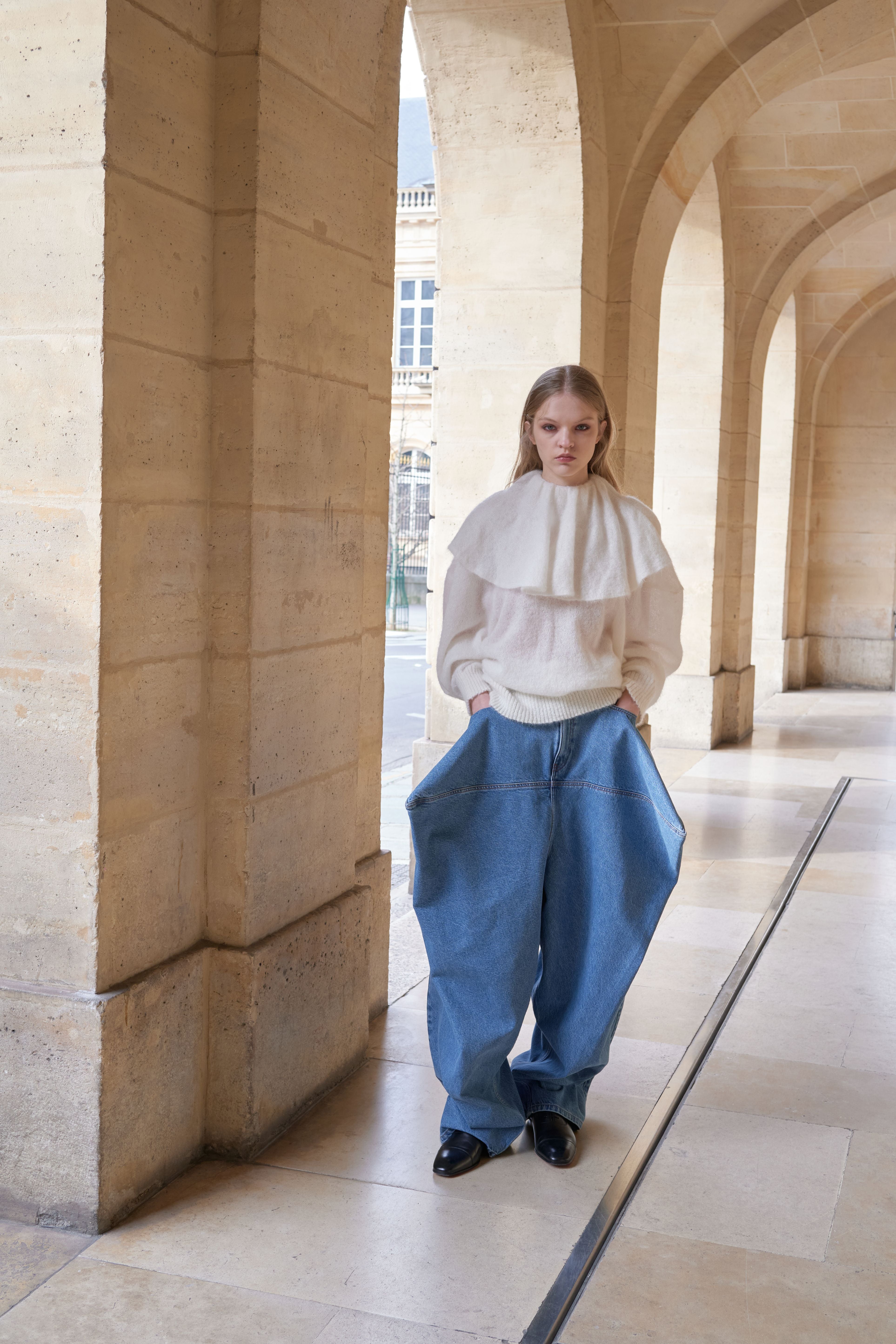Person wearing a white top and blue pants standing in an architectural setting with stone walls and columns.