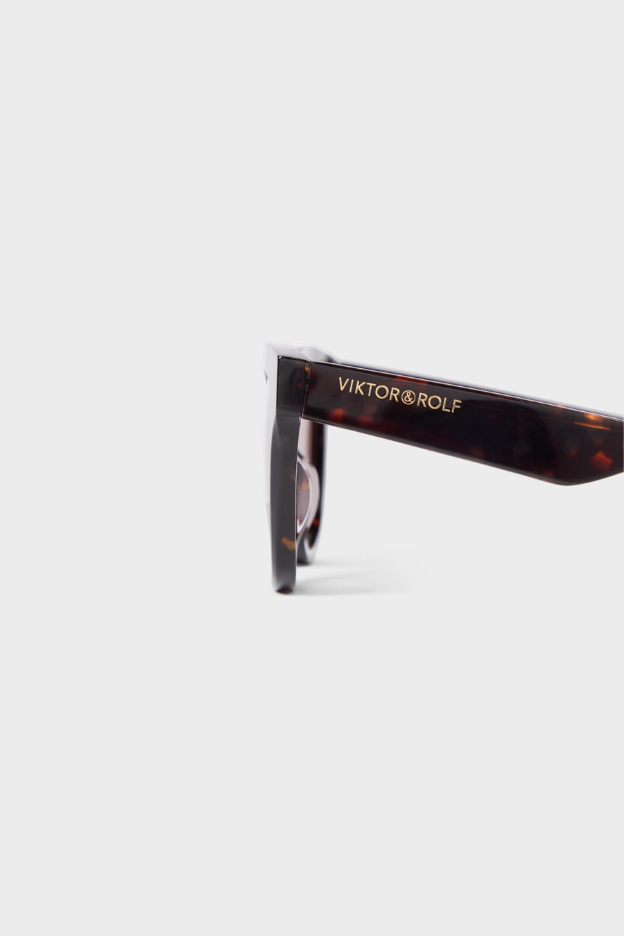 Tortoiseshell sunglasses arm featuring a sleek design with the brand logo in gold lettering against a light solid background.